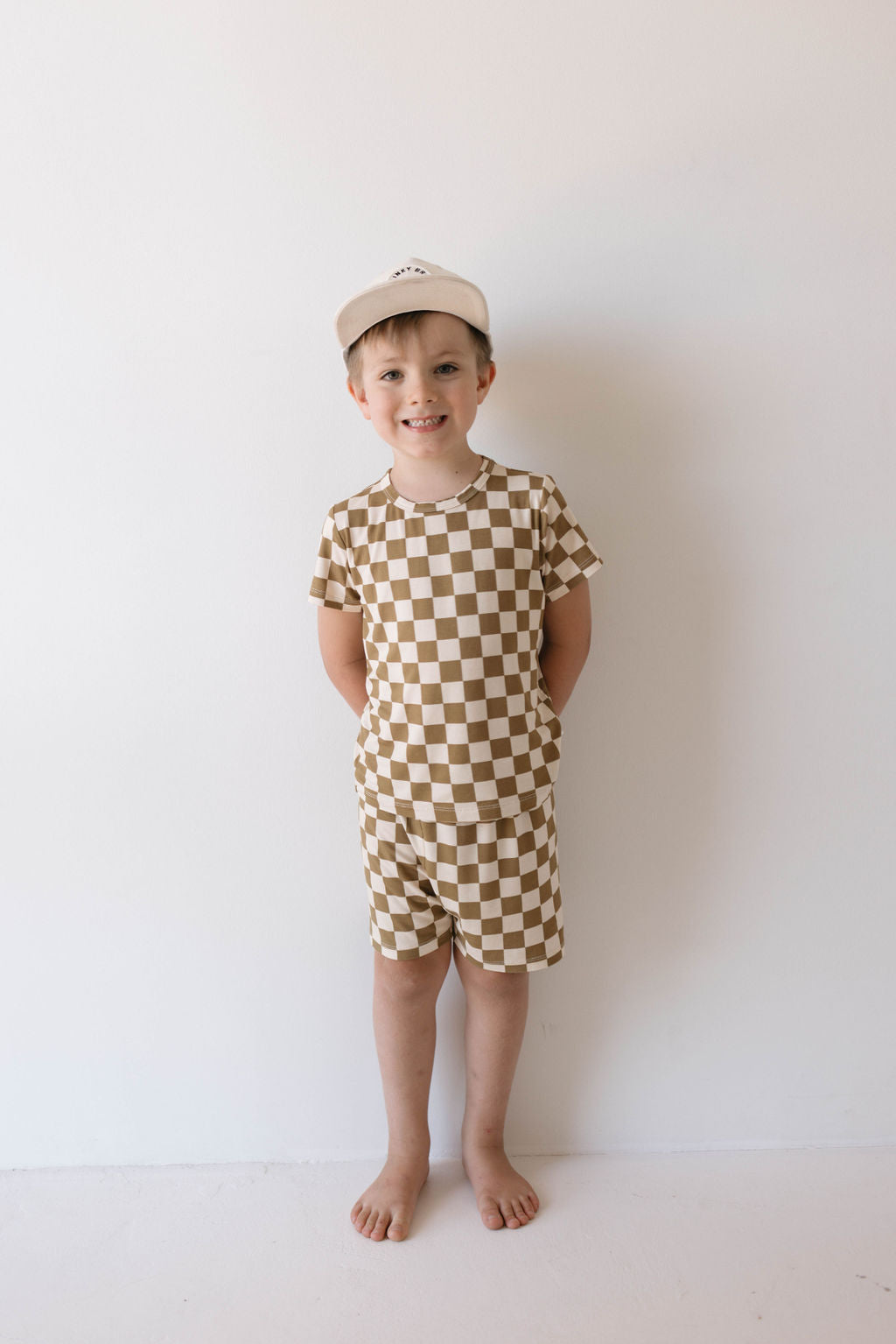 A young boy stands against a plain white background, smiling. He is wearing the Bamboo Short Set in Olive Checkerboard from forever french baby, along with a beige baseball cap. His hands are behind his back and he is barefoot, embodying the comfort of hypo-allergenic clothing for carefree play.