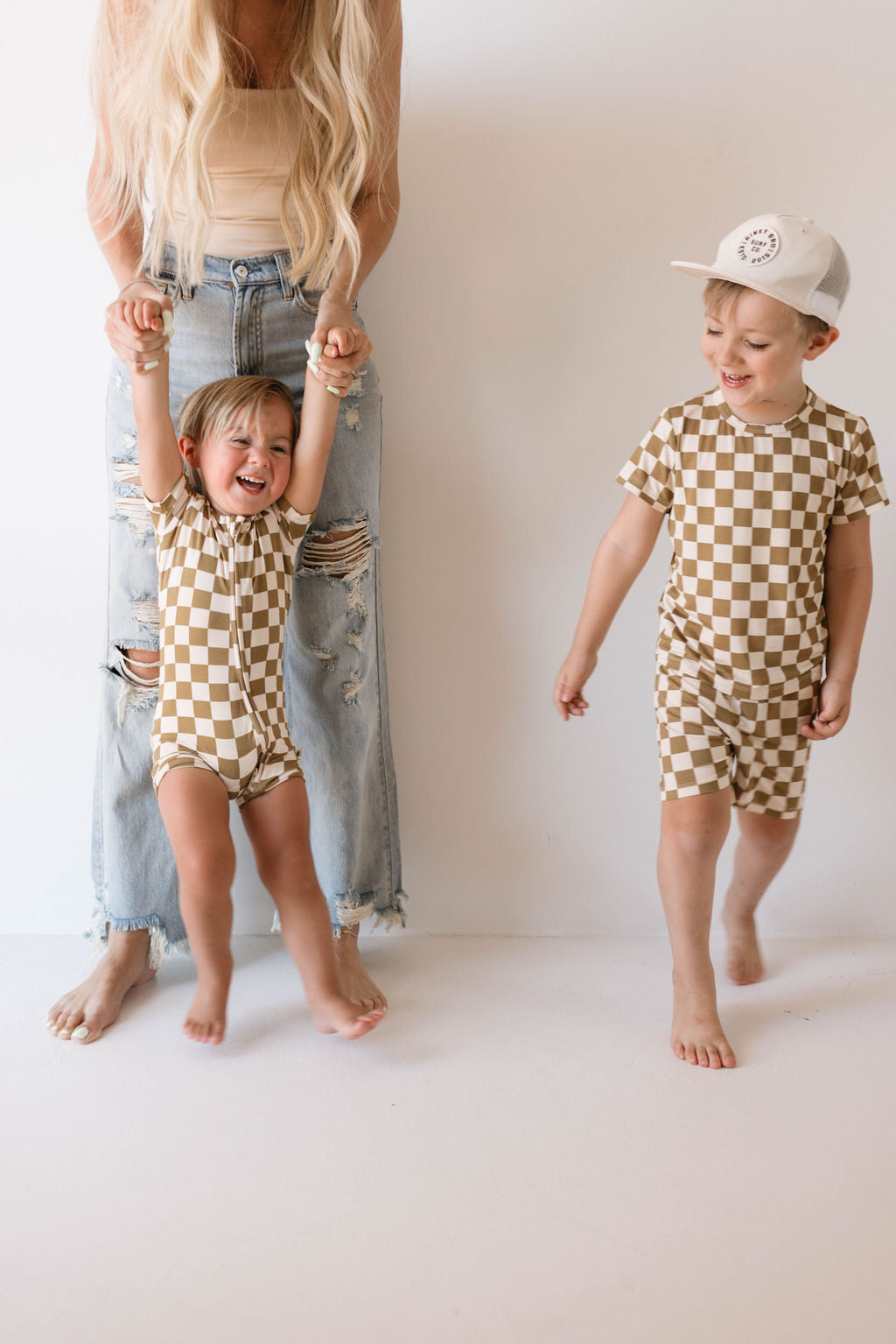 An adult, visible from the waist down and dressed in distressed jeans, holds the hands of a laughing child who is wearing a breathable "Bamboo Short Set" in olive checkerboard pattern by forever french baby. Another child in matching hypo-allergenic clothing walks barefoot nearby while wearing a white cap.
