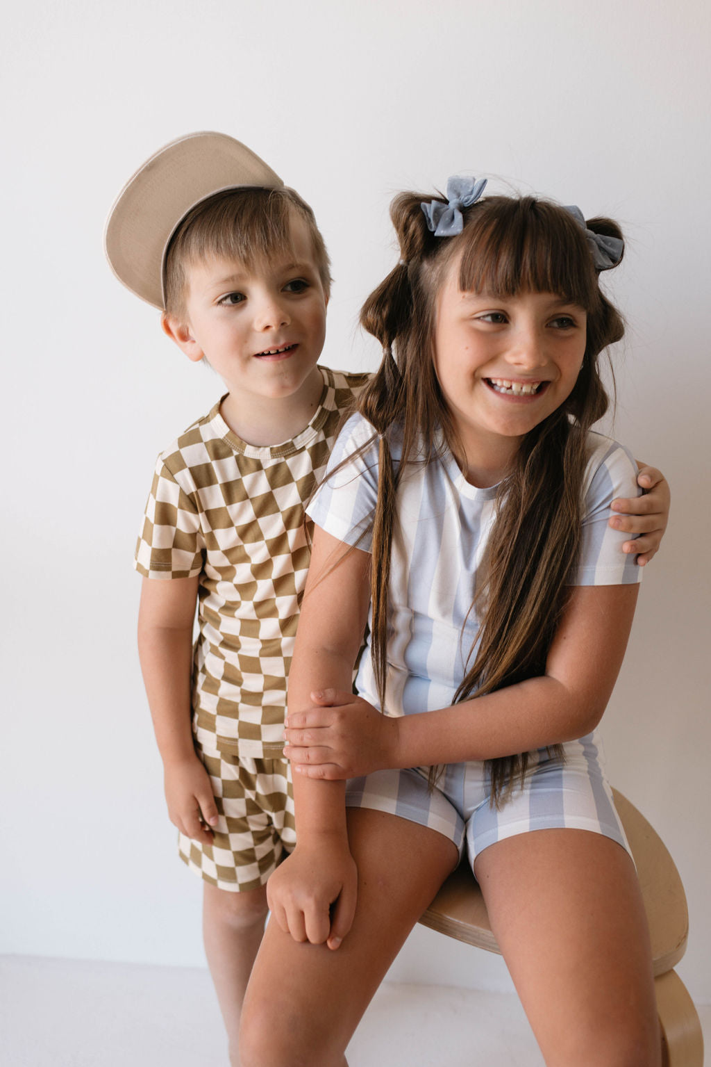Two children pose against a plain background. The boy on the left wears forever french baby's Bamboo Short Set in Olive Checkerboard paired with a backwards cap, smiling with his arm around the girl's shoulder. The girl on the right is dressed in breathable checkered babywear in blue and white, smiling with long hair adorned with bows.