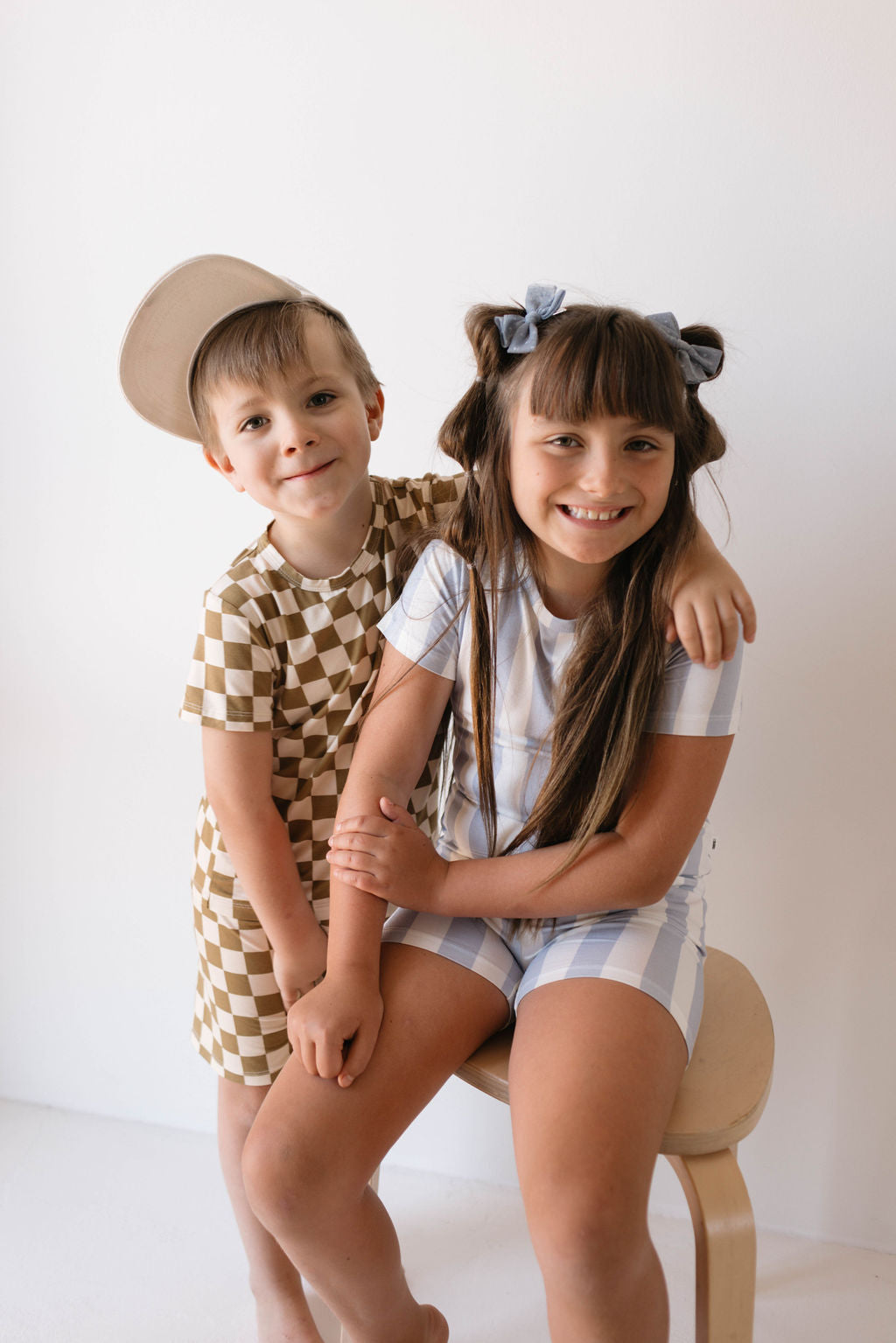 Two smiling children share a friendly moment. The child on the left, dressed in Forever French Baby's Bamboo Short Set in Olive Checkerboard and a beige cap, enjoys the comfort of breathable babywear. The child on the right, seated on a stool, wears a light blue and white checkered outfit with hair styled in two pigtails.