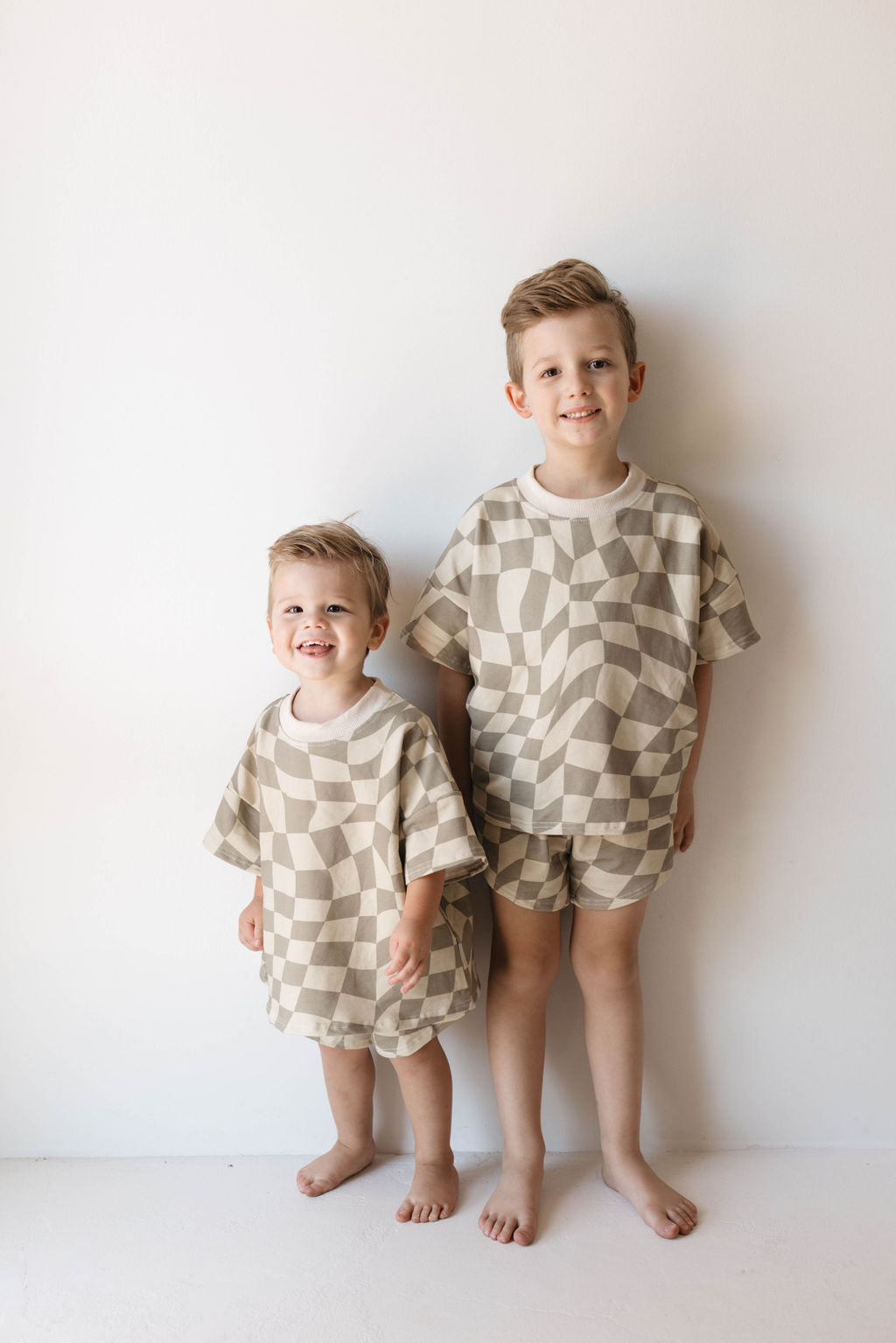 Two young children stand barefoot against a plain white wall, both smiling in matching Short Set | Wavy Checkerboard outfits by forever french baby. The older child stands straight while the younger one leans forward with an animated expression, showcasing their trendy children's clothing made from organic cotton.