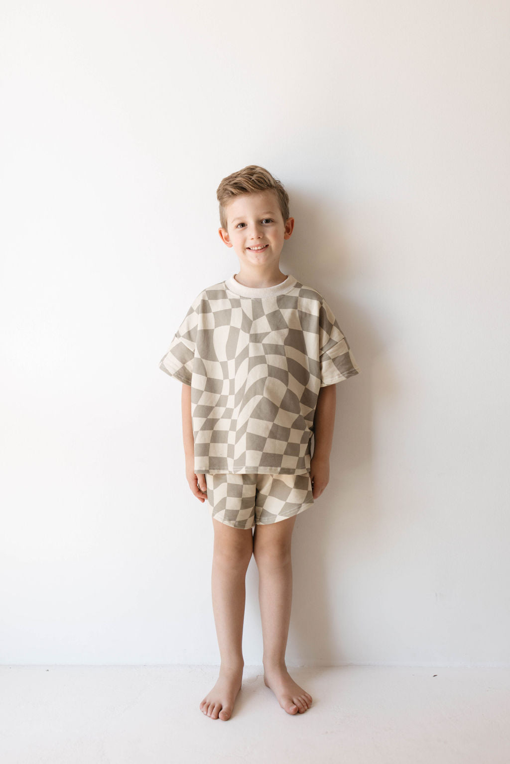 A young boy stands barefoot against a white background, smiling at the camera. He is dressed in the forever french baby Short Set | Wavy Checkerboard, made from organic cotton. The set includes a loose, short-sleeved shirt and shorts featuring a neutral-toned checkerboard pattern. This trendy children's clothing exudes effortless charm.
