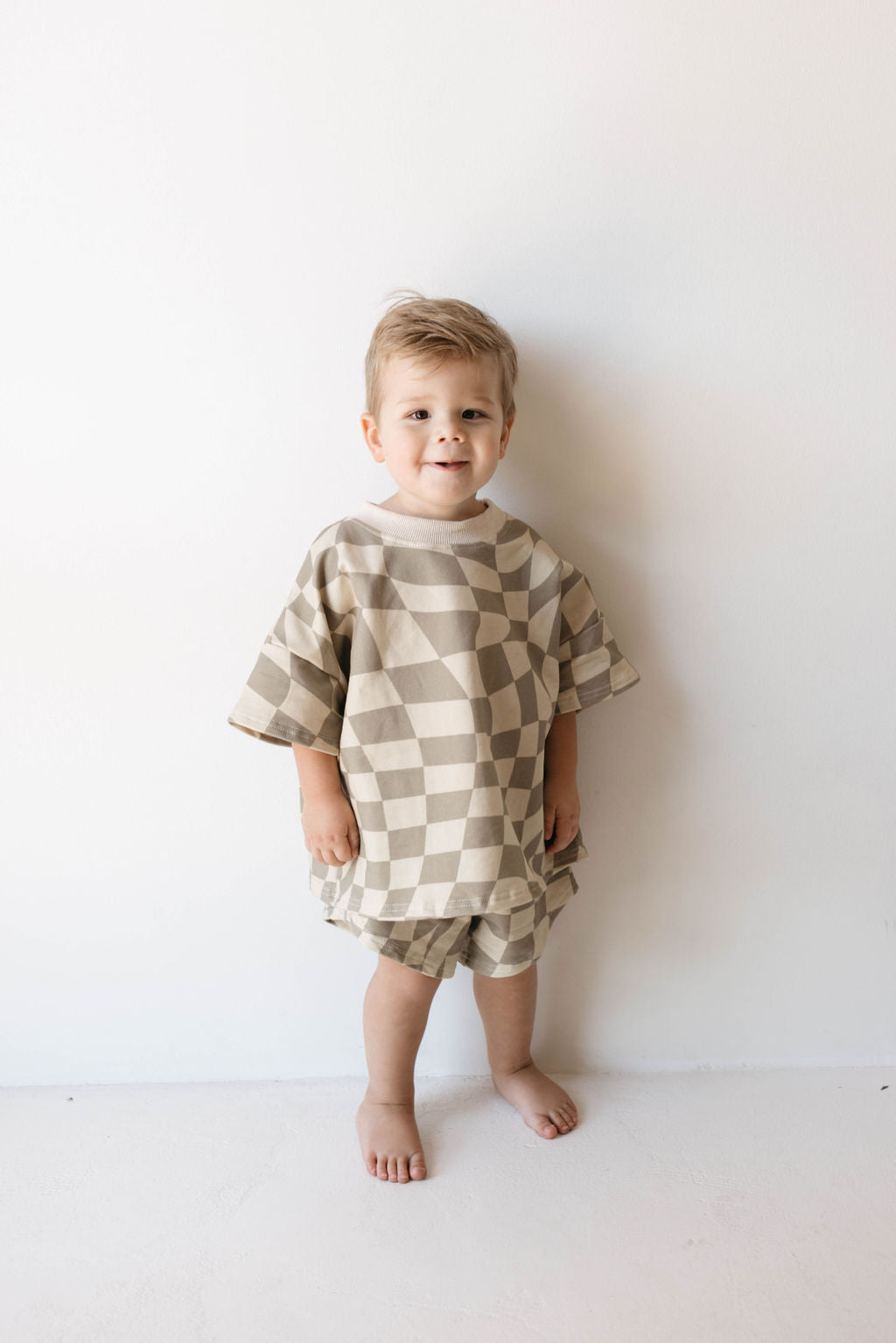 A young child stands barefoot against a plain white wall, dressed in the trendy, oversized "Short Set | Wavy Checkerboard" from forever french baby. The organic cotton ensemble, featuring a beige and white checkered pattern, perfectly complements the child's short light brown hair and slight smile.