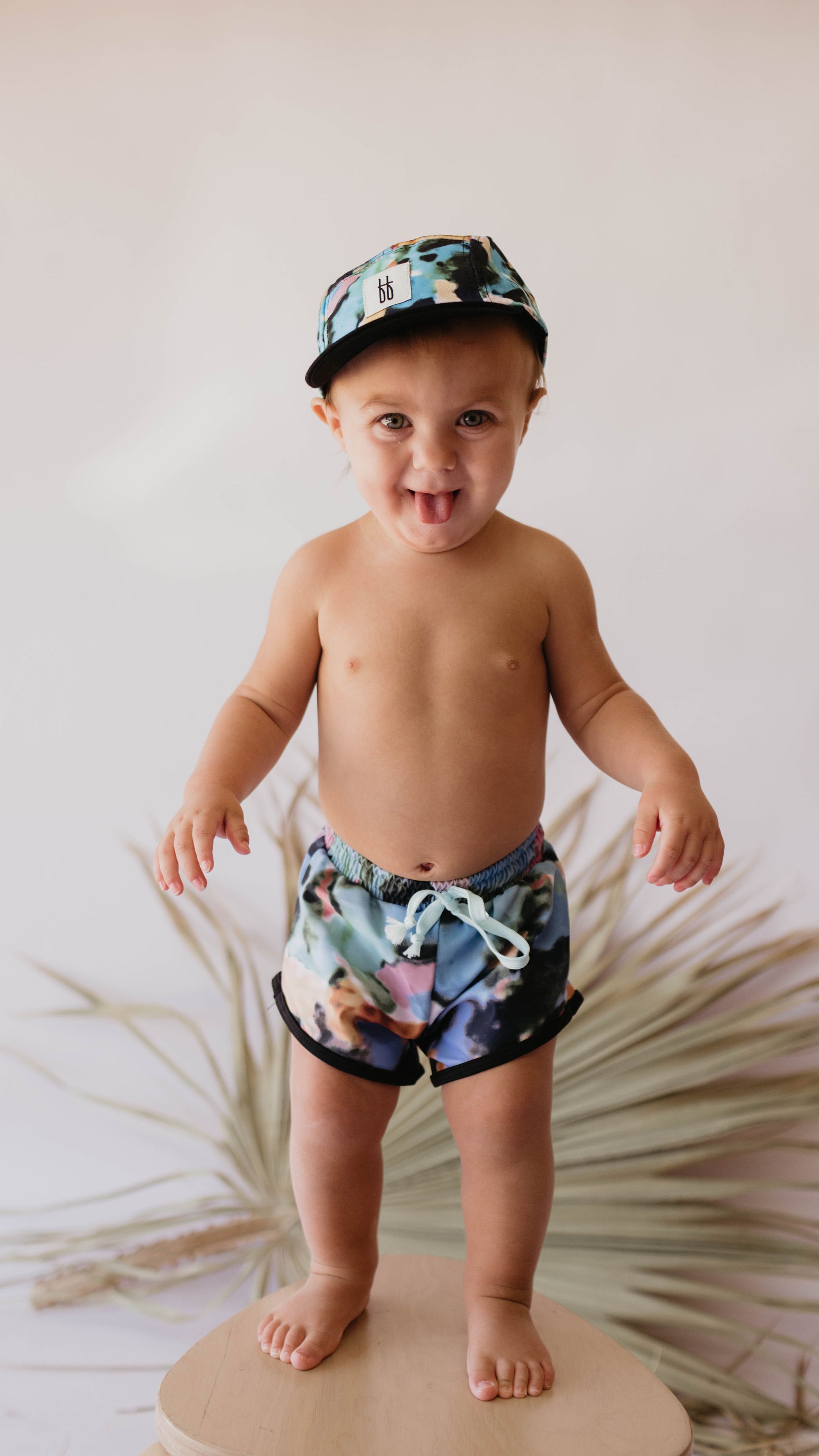A joyful baby confidently stands on a light-colored surface, wearing the Snap Back | Charli Print cap and matching shorts with a drawstring from forever french baby. The delightful scene resembles an exclusive watercolor print, set against a blurred, neutral-toned background featuring spiky plant leaves. The baby looks happy and playful.