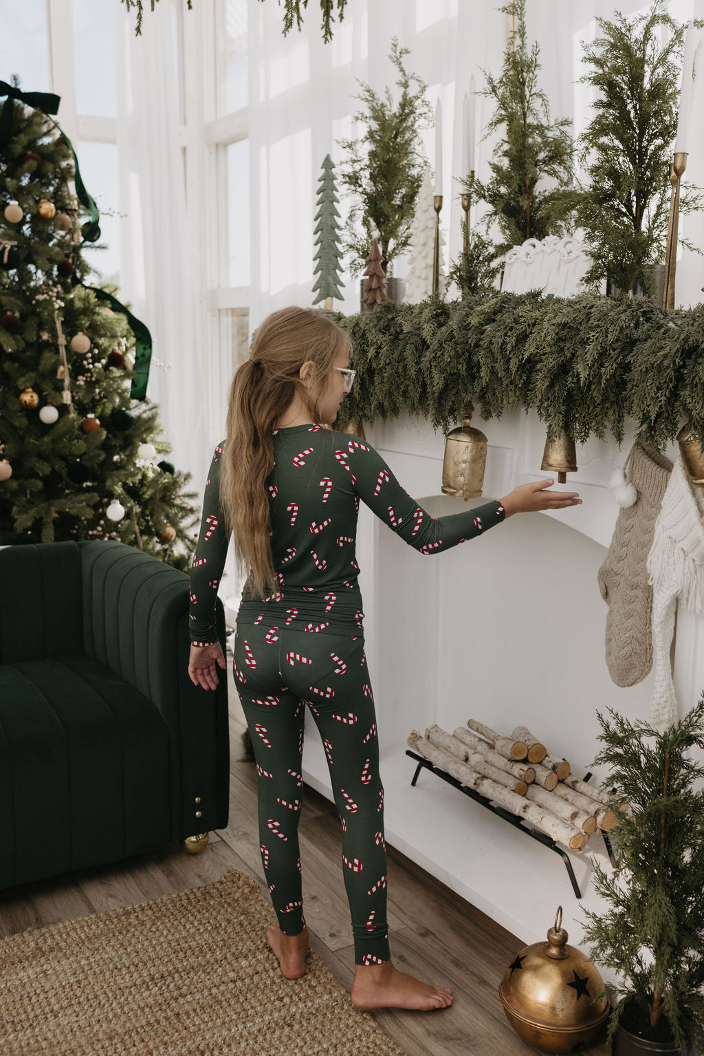 A girl wearing the Bamboo Two Piece Pajamas from Candy Cane Lane by forever french baby decorates a mantel with evergreen garlands and hanging stockings. A Christmas tree adorned with ornaments stands nearby in the cozy room.