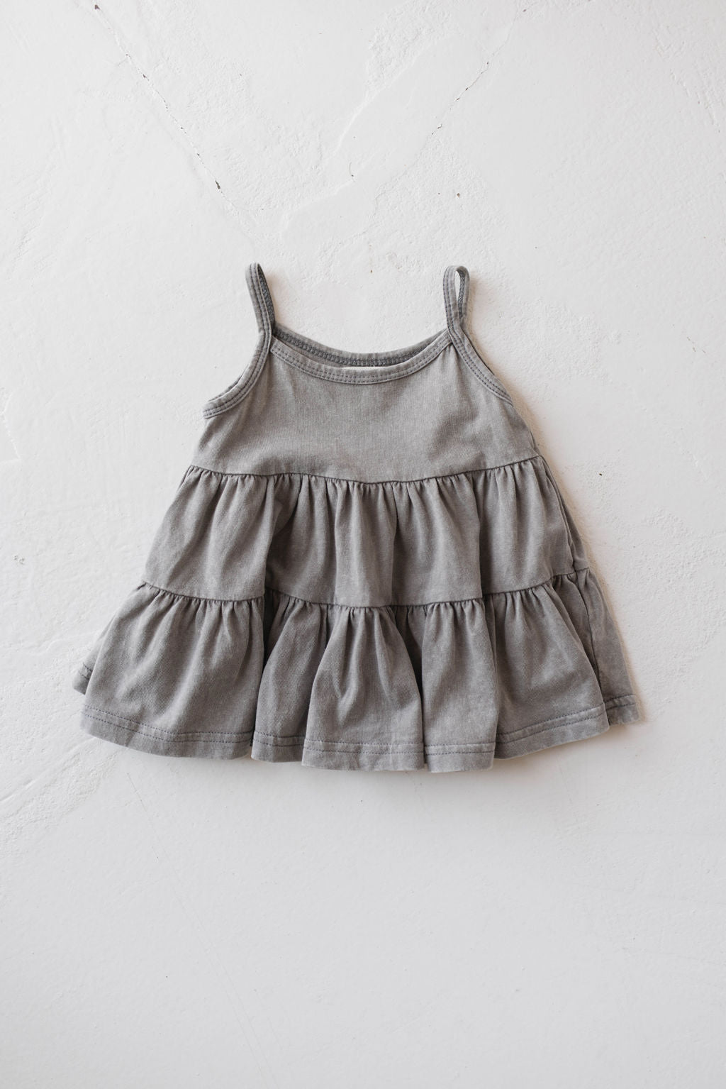 The Children's Dress | Vintage Washed Steel from forever french baby, in a small size, lies flat on a white textured surface. The sleeveless dress features a tiered, ruffled design and thin shoulder straps.