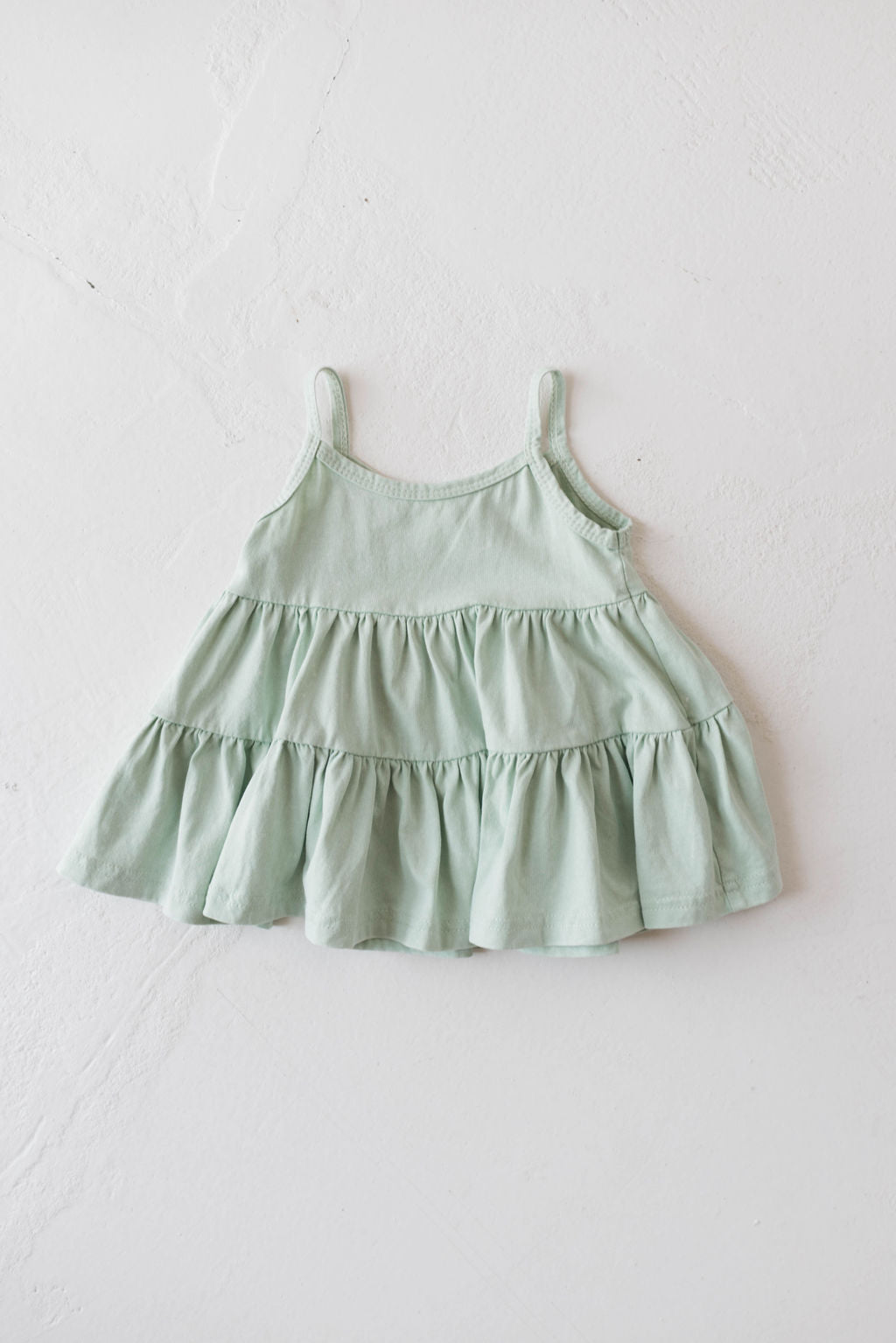 The Children's Dress in Vintage Washed Spearmint by forever french baby, featuring thin straps and a three-tiered ruffle design, is laid flat on a white textured surface.