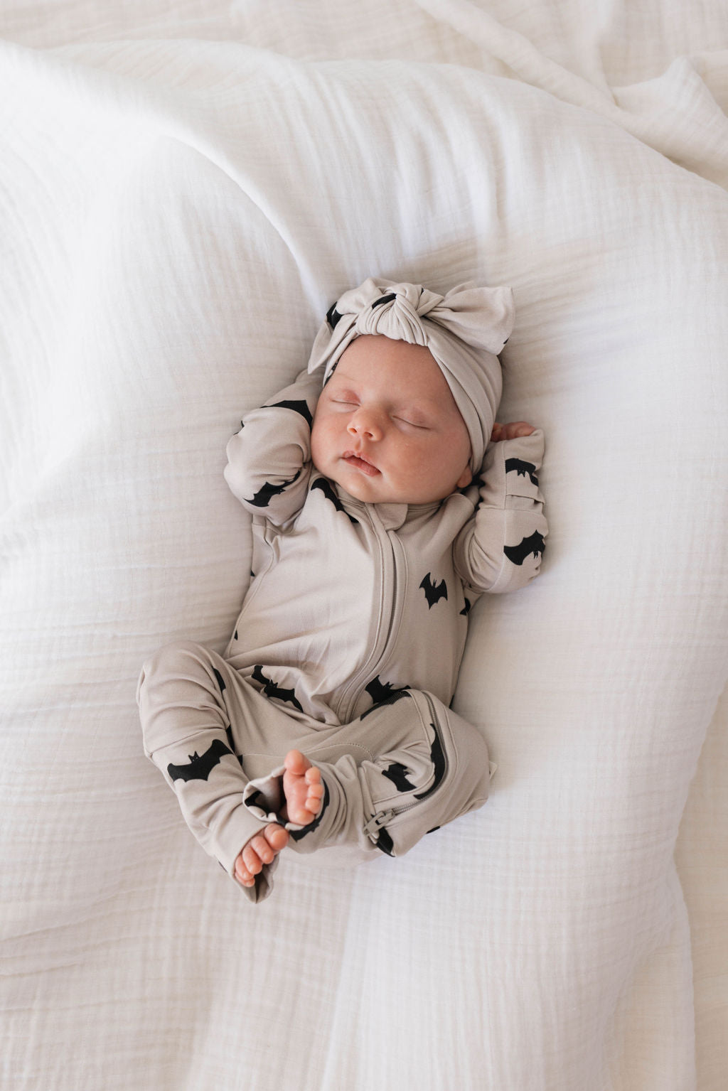 A newborn baby sleeps peacefully on a soft, white blanket. The baby, dressed in a light gray onesie with black bat patterns made from bamboo material, and an adjustable Bamboo Head Wrap | It's Bats! by forever french baby with a knotted bow, has their hands positioned near their head and their legs curled up.