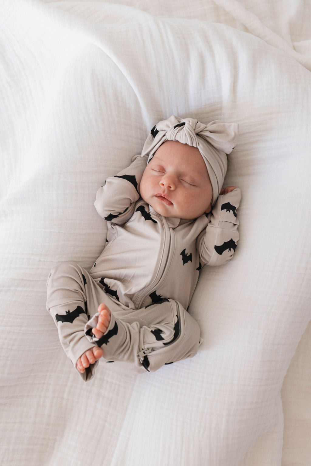A sleeping baby lies on a white blanket, dressed in forever french baby's Bamboo Zip Pajamas | It's Bats!, featuring light beige and black bat patterns. The baby wears a matching headband with a large bow and has their arms raised near their head.