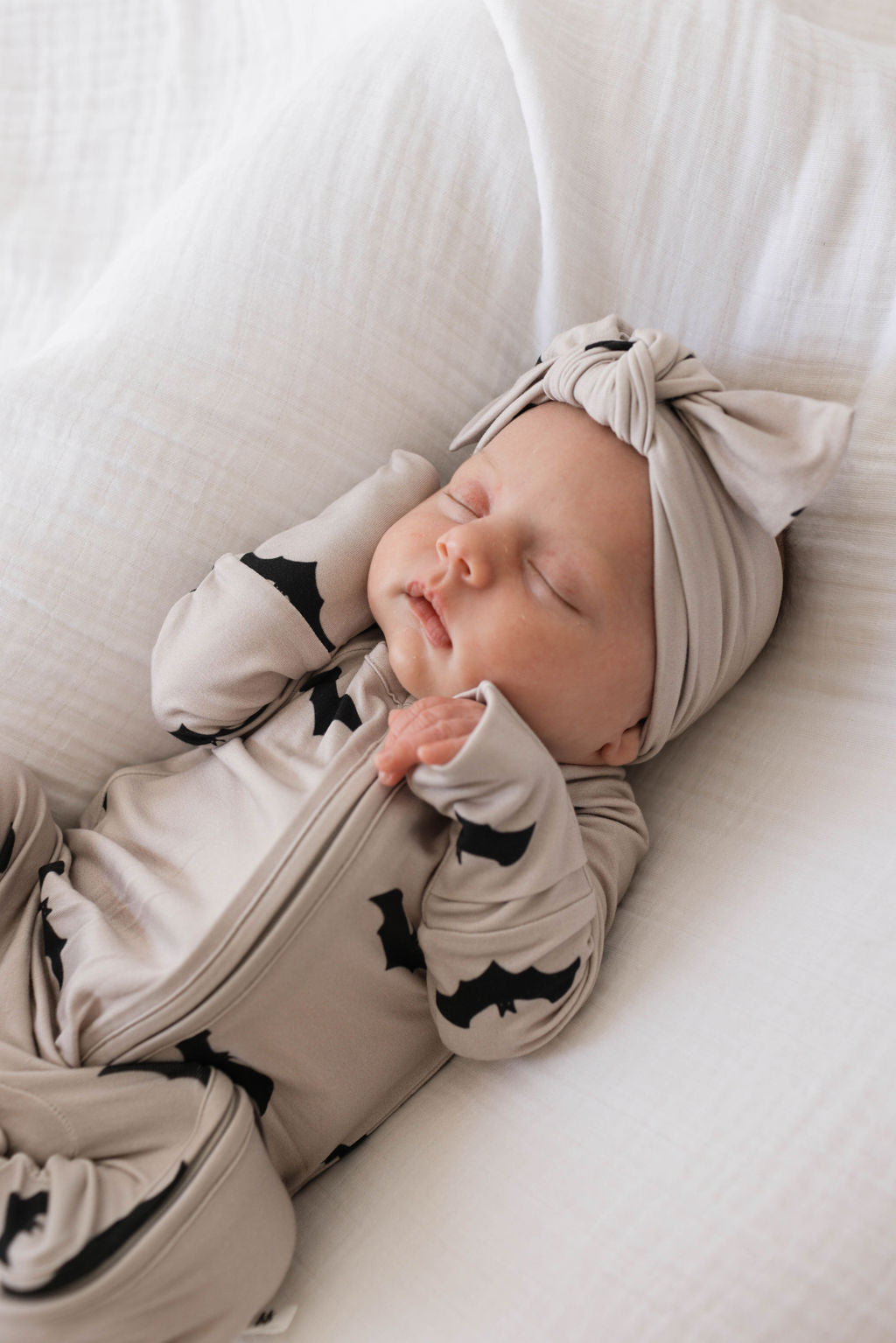 A baby is peacefully sleeping on a white blanket. The infant is wearing forever french baby's Bamboo Zip Pajamas | It's Bats!, adorned with dark bat patterns and has a matching headband with a bow. The hypo-allergenic bamboo fabric ensures comfort as the baby's arms are raised beside its head.