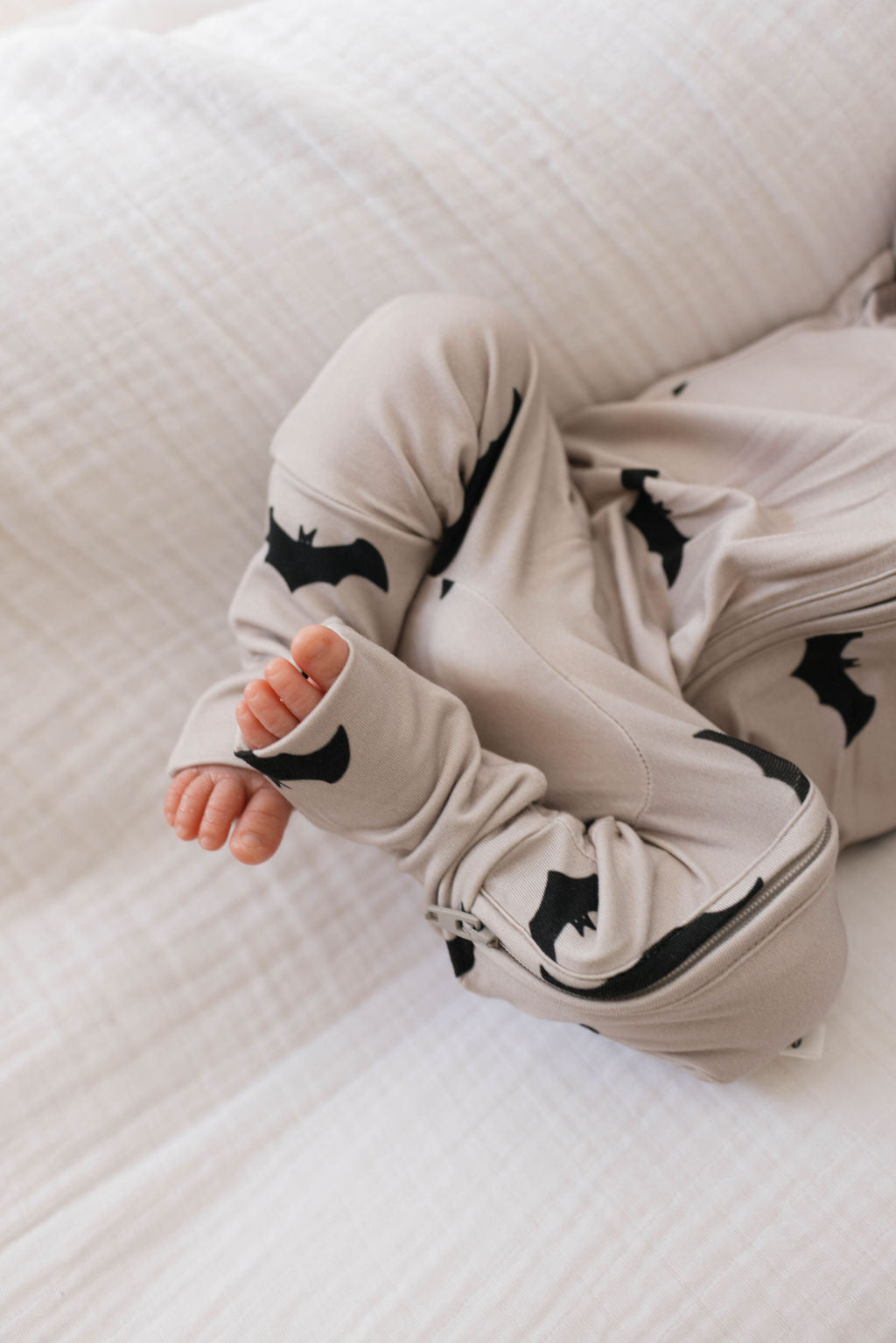 A baby in Forever French Baby's Bamboo Zip Pajamas | It's Bats! with black bat patterns lies on a white quilted blanket. The baby's hands and tiny feet are visible, with their legs slightly bent and arms relaxed.