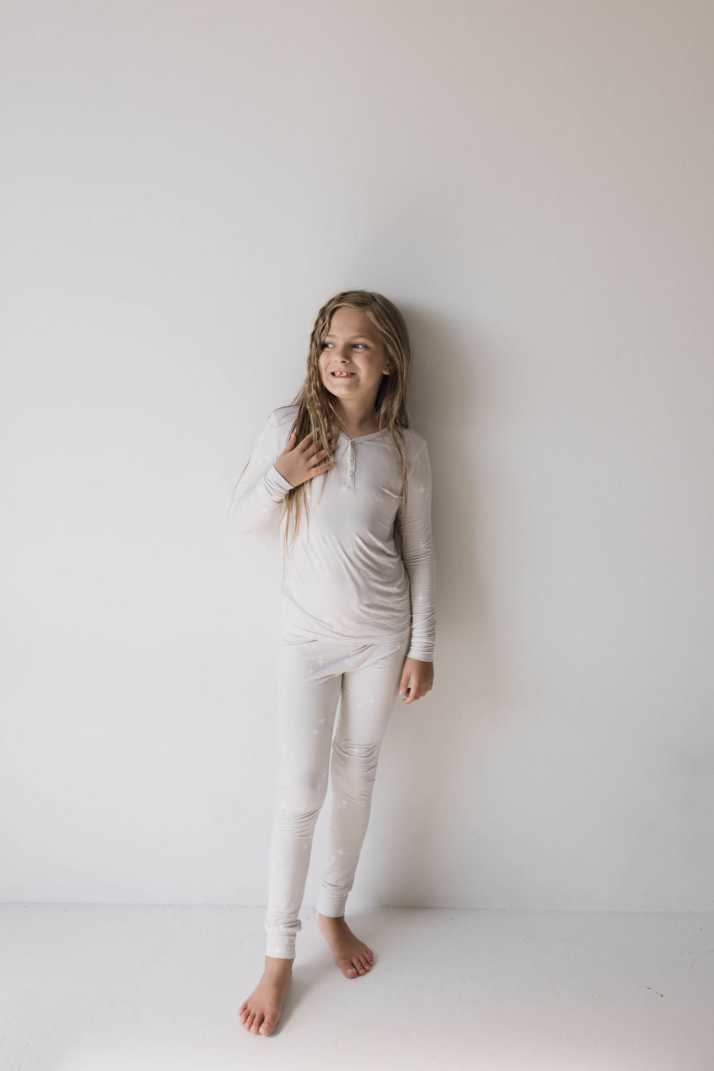 A young girl with long hair stands against a white wall, smiling and looking off to her left. She is wearing Forever French Baby Bamboo Two Piece Pajamas in the Sparkle! design that are form-fitting and long-sleeved, and she is barefoot. The lighting is soft, giving the scene a calm and cheerful mood.