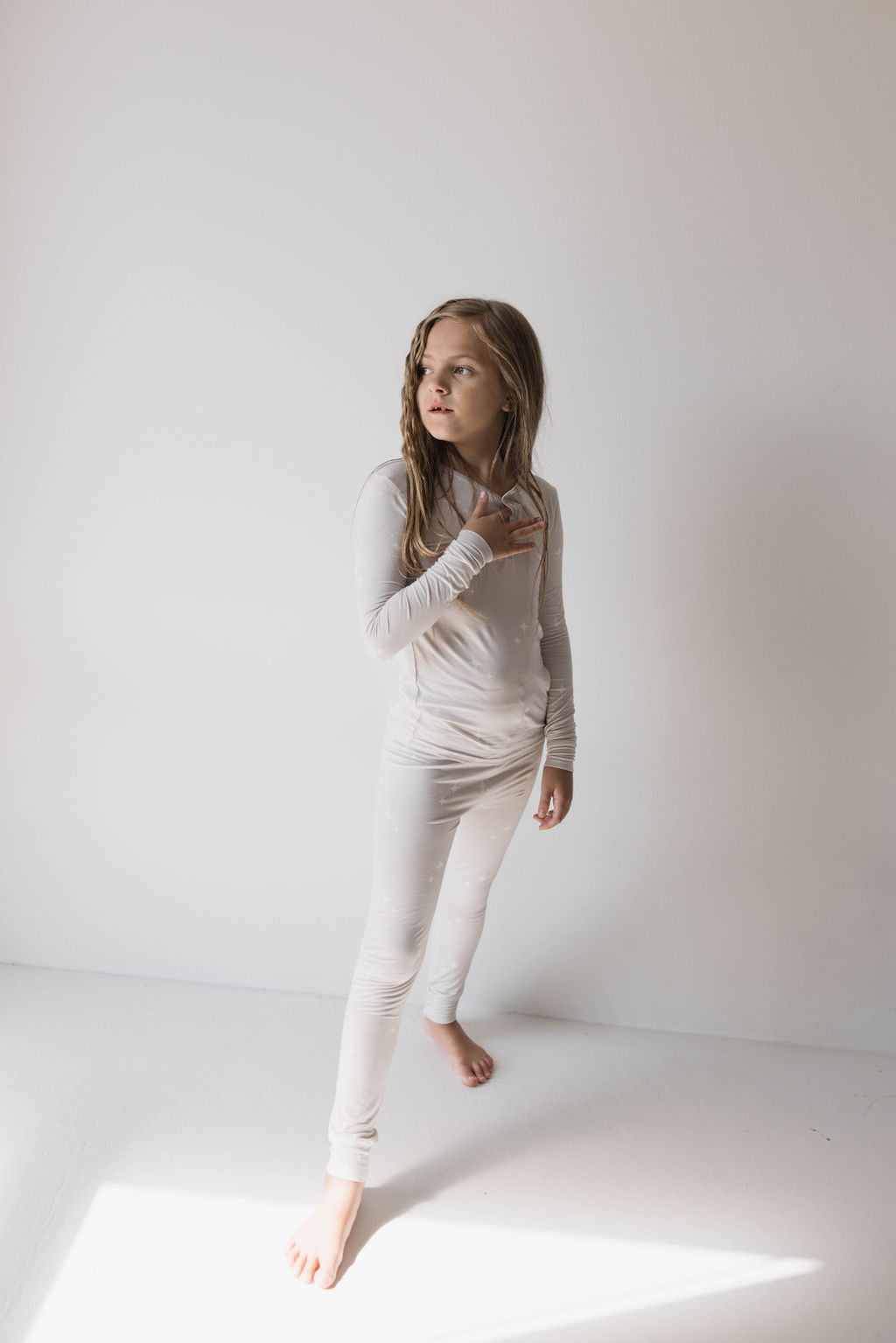 A young girl with long hair stands barefoot on a white floor against a plain white wall, wearing Forever French Baby's Bamboo Two Piece Pajamas in the "Sparkle!" design. She looks off to the side with one hand slightly raised near her chest.
