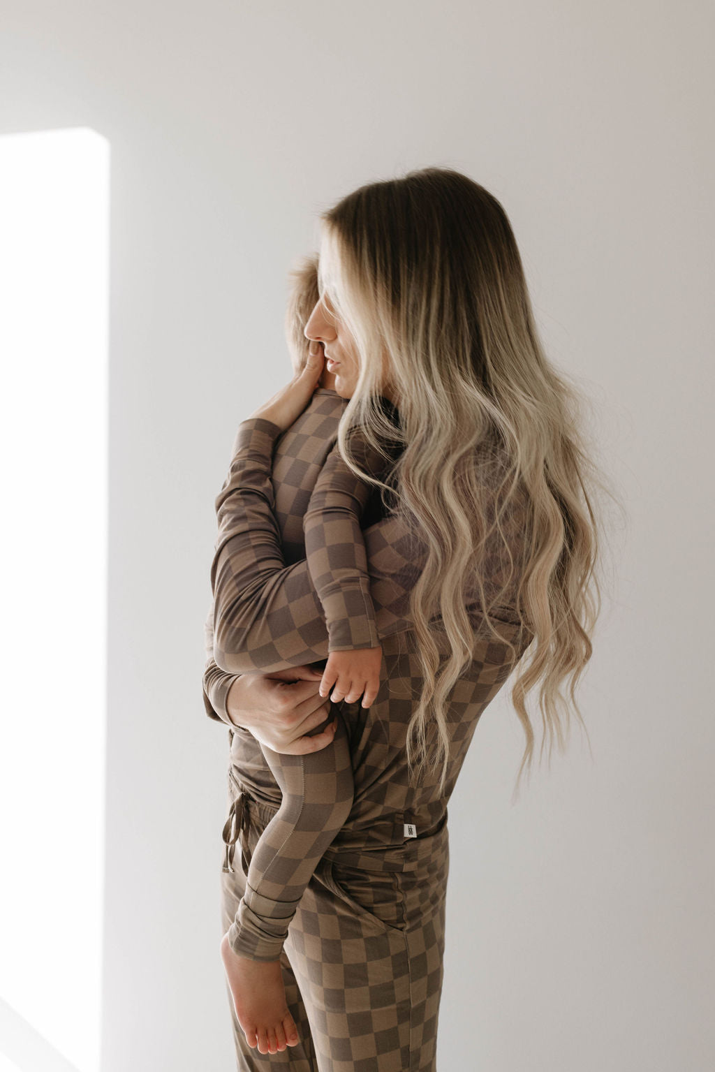 A woman with long, wavy, blonde hair holds a child in a gentle embrace. They are both dressed in matching forever french baby Women's Bamboo Pajamas featuring a Faded Brown Checkerboard pattern, made from hypo-allergenic fabric. The scene is set against a plain white background with soft light illuminating them from the left.