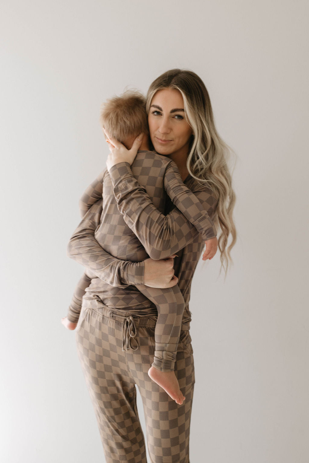 A woman with long, wavy blond hair is holding a small child dressed in matching checkered pajamas from the forever french baby brand. Both are wearing Women's Bamboo Pajamas in Faded Brown Checkerboard, made from hypo-allergenic materials. She is looking at the camera with a neutral expression while the child rests their head on her shoulder. The background is plain and light-colored.