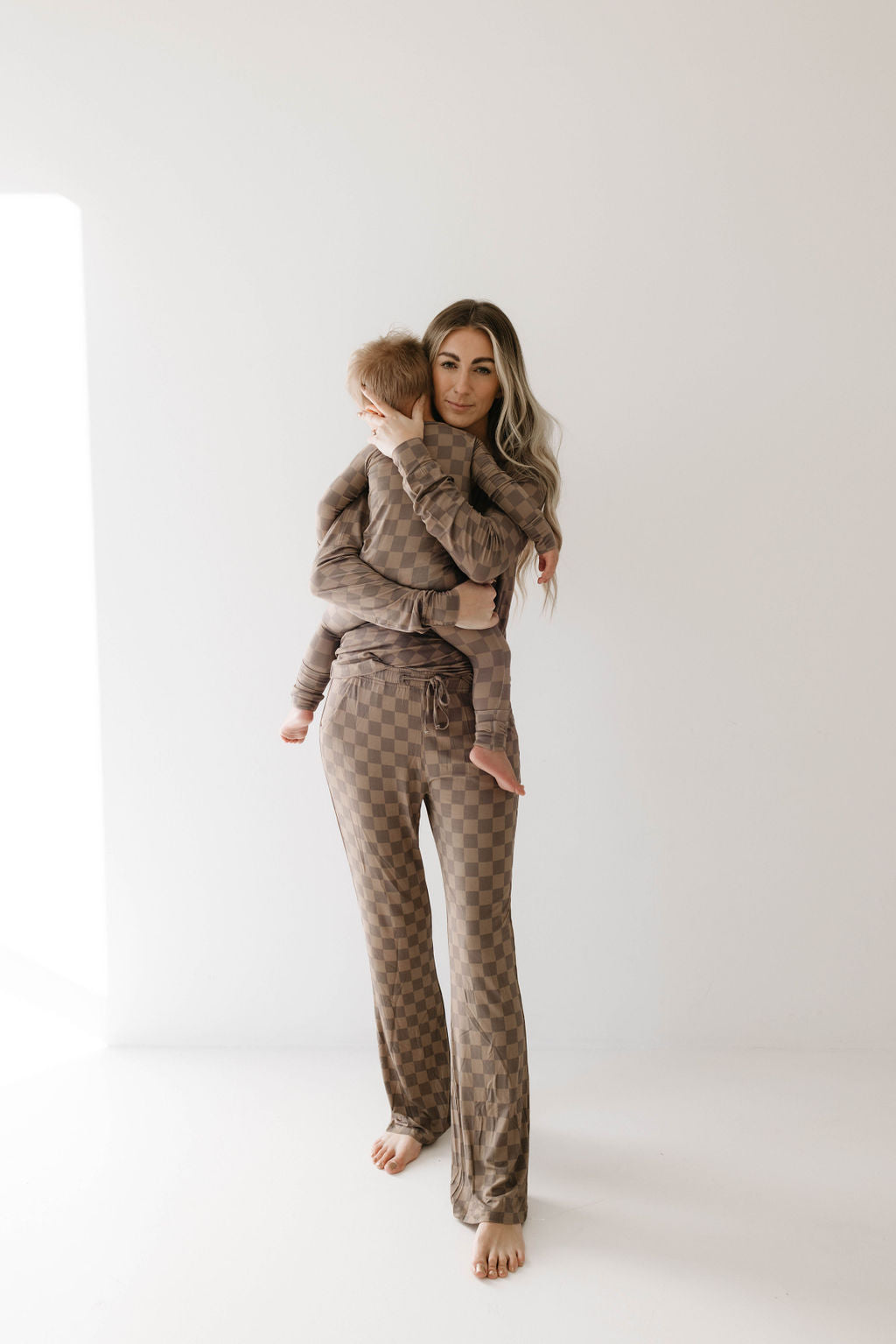 A woman with long, wavy hair stands barefoot, holding a young child. Both are wearing matching outfits: Women’s Bamboo Pajamas in Faded Brown Checkerboard from forever french baby. They are positioned against a minimal, light-colored background with natural light streaming in from the left.