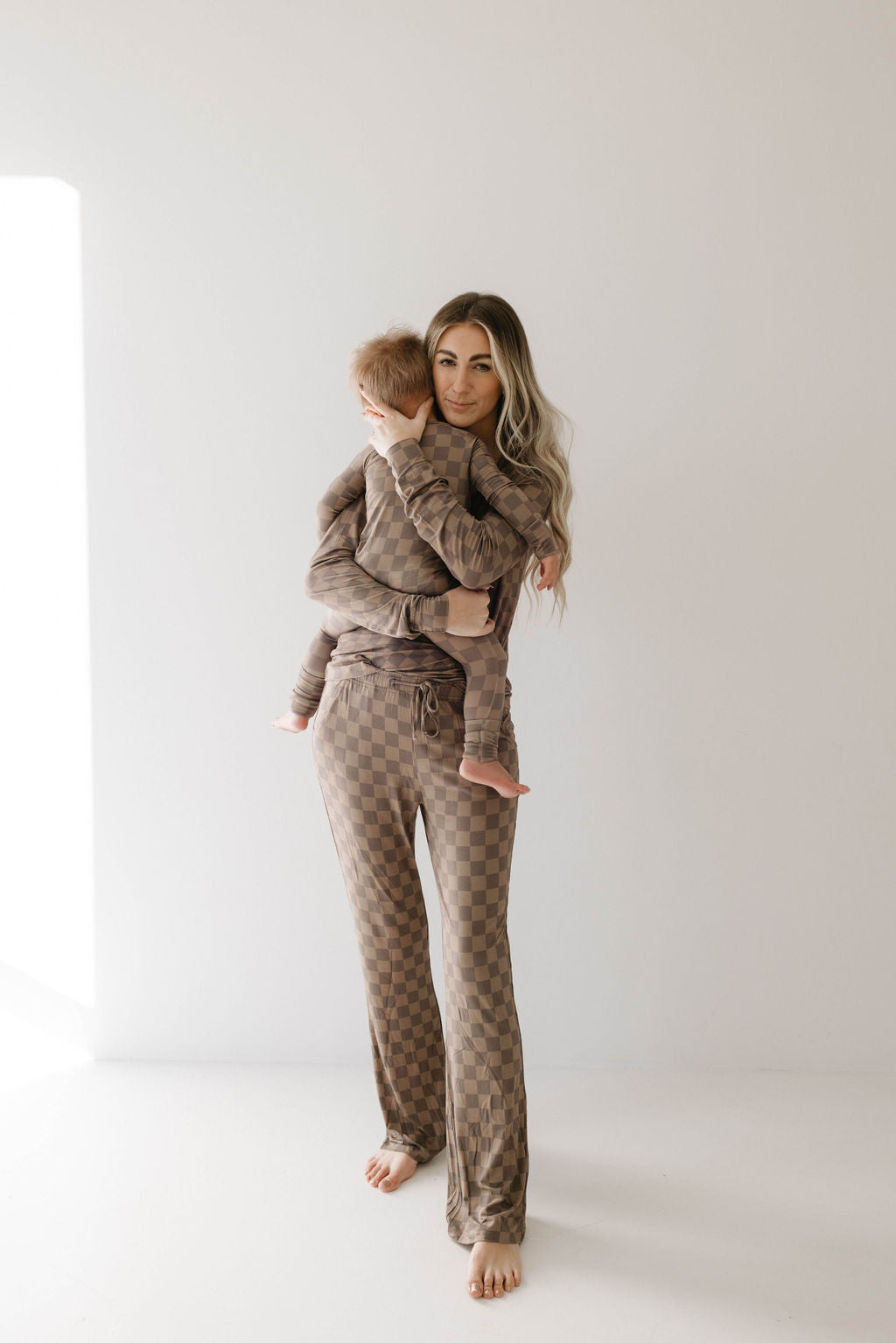 A woman with long, wavy hair stands barefoot in a minimalist room wearing forever french baby's Women's Bamboo Pajamas in Faded Brown Checkerboard, holding a small child dressed in a matching outfit. The child rests their head on the woman's shoulder.