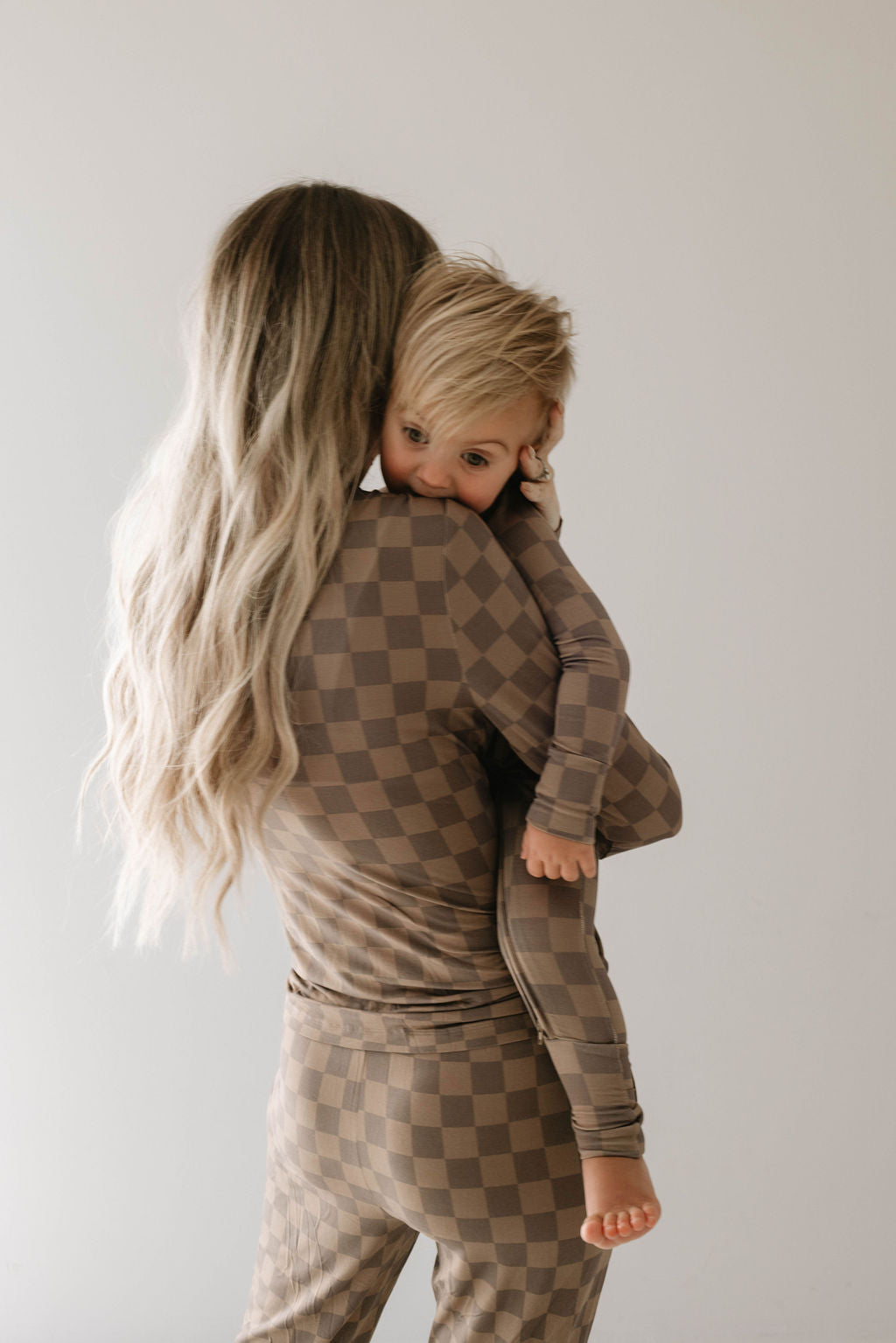 A woman with long, wavy blonde hair, seen from behind, is holding a small child who is hugging her shoulder. Both are wearing matching Women's Bamboo Pajamas in Faded Brown Checkerboard by forever french baby. The child looks towards the camera with a calm expression while the woman faces away.
