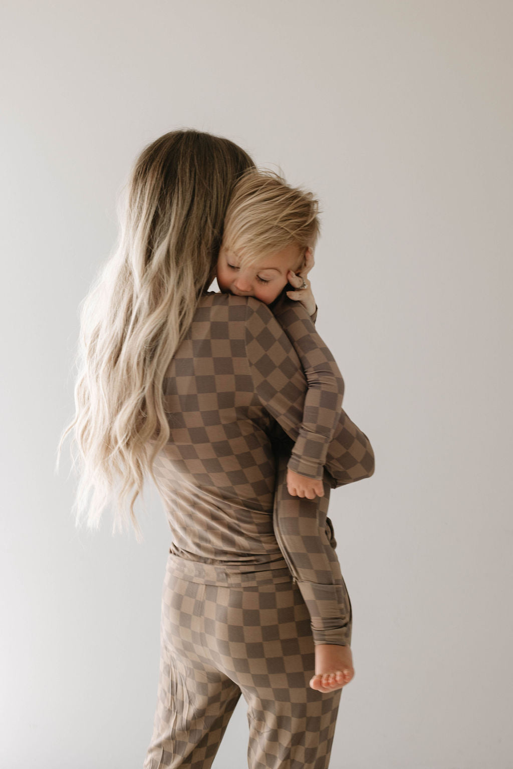 A woman with long, wavy blonde hair cradles a sleeping toddler on her shoulder. Both are dressed in matching hypo-allergenic bamboo pajamas from forever french baby, featuring a faded brown checkerboard pattern. The background is plain and light-colored.