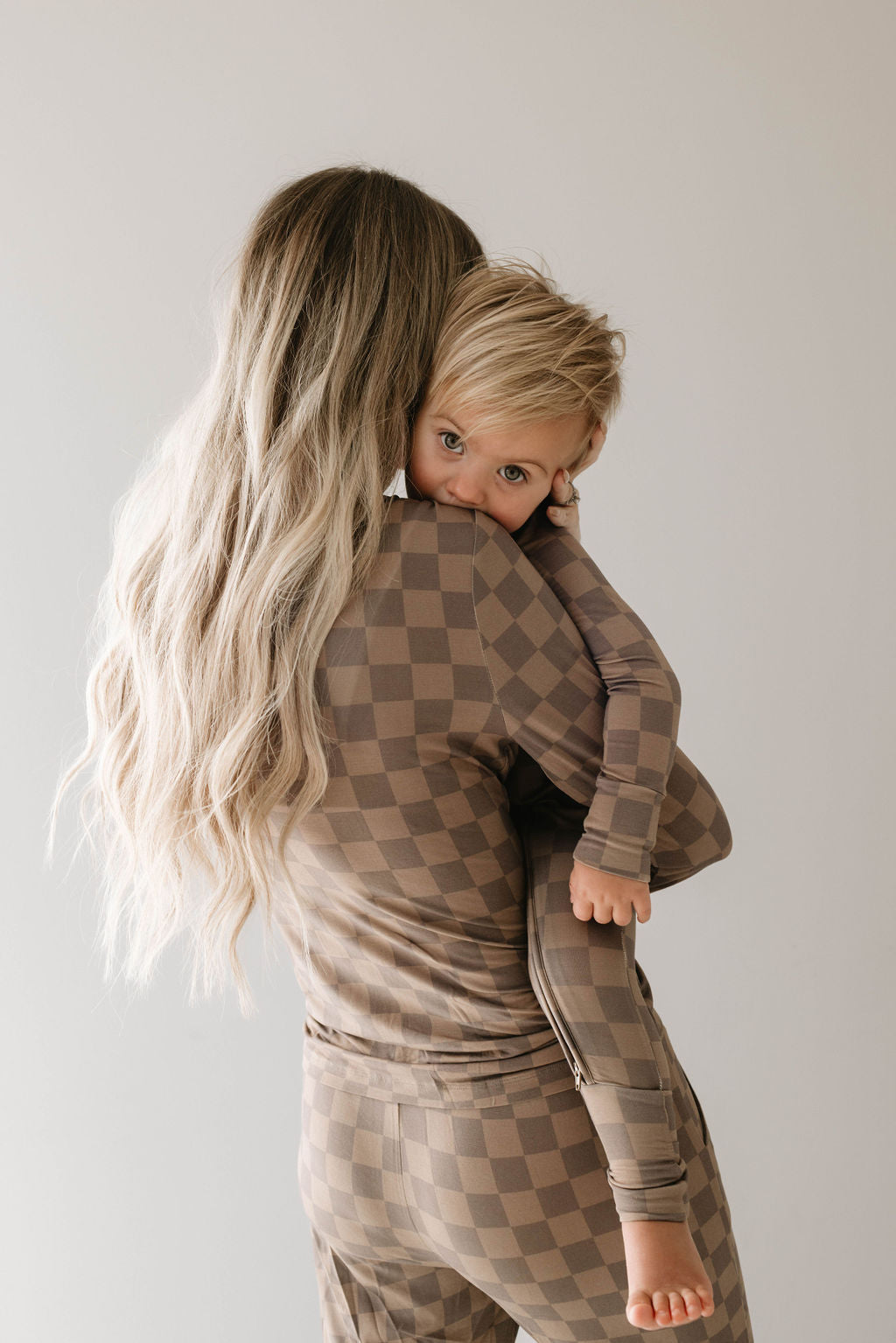 A woman with long, blonde hair, wearing forever french baby's hypo-allergenic Women's Bamboo Pajamas in Faded Brown Checkerboard, holds a young child dressed in matching pajamas. The child looks directly at the camera while being carried on the woman's hip, leaning their head on her shoulder.