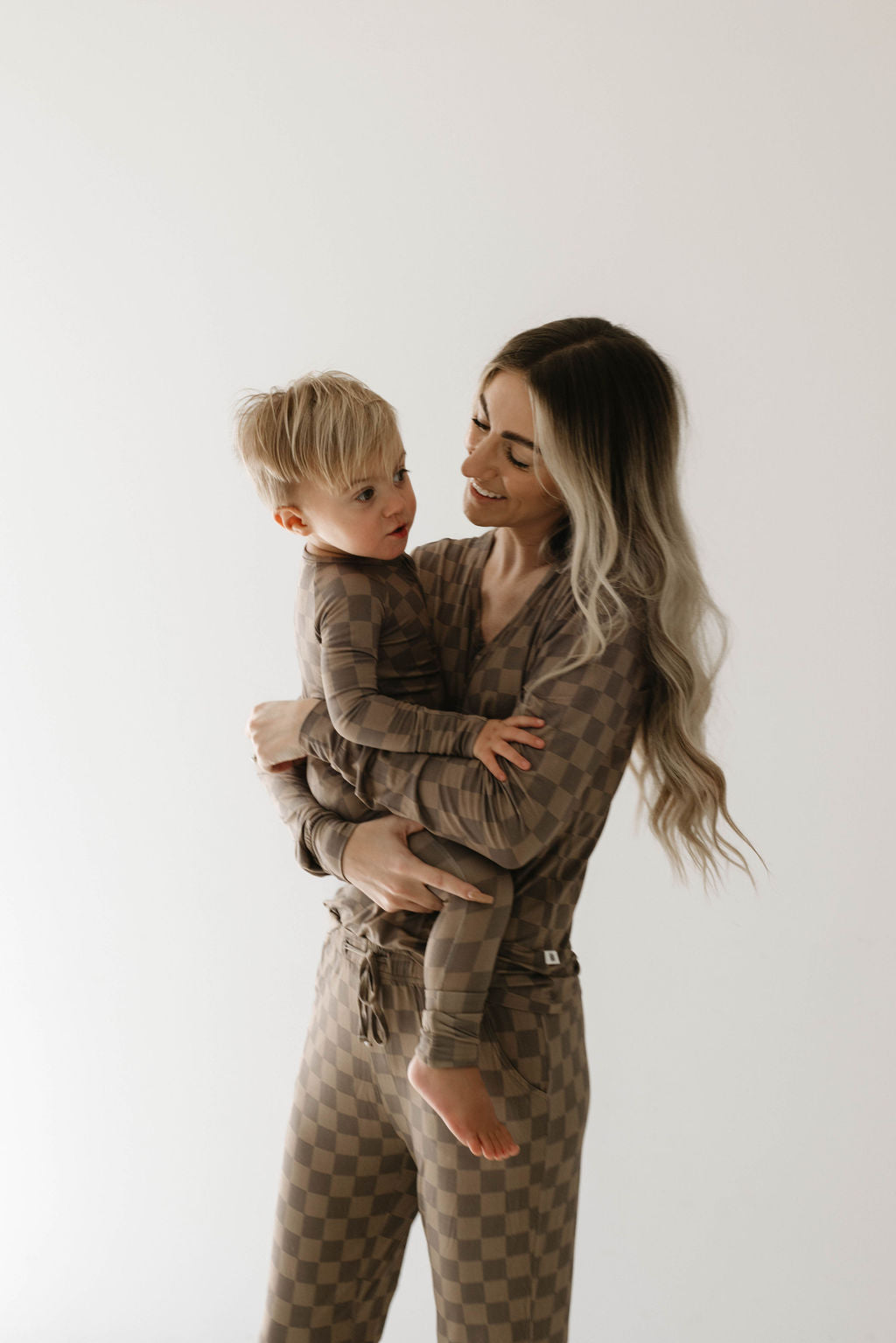 A woman with long, wavy hair holds a young child in her arms. Both are wearing matching, breastfeeding accessible outfits in a neutral color, specifically the Women's Bamboo Pajamas in Faded Brown Checkerboard by forever french baby. They are facing each other and smiling, standing against a plain, light-colored background.