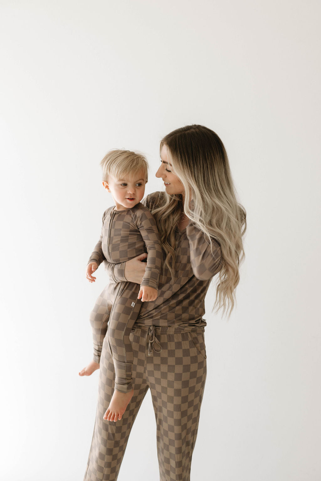A woman with long, light blonde hair is holding a small child. Both are wearing matching Women's Bamboo Pajamas in Faded Brown Checkerboard, a stylish set from forever french baby made from hypo-allergenic fabric. They stand against a plain, light background, looking at each other with smiles.