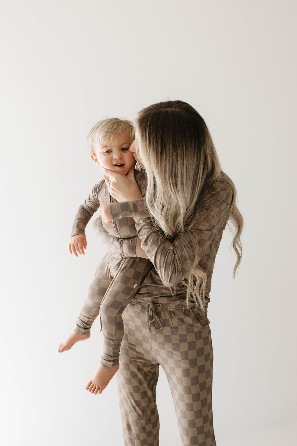 A woman with long, wavy hair dressed in forever french baby's Women's Bamboo Pajamas in Faded Brown Checkerboard is holding a smiling blond toddler wearing a matching outfit. They are standing against a plain, light-colored background. The woman is looking at the child affectionately in their hypo-allergenic, breastfeeding accessible pajamas.