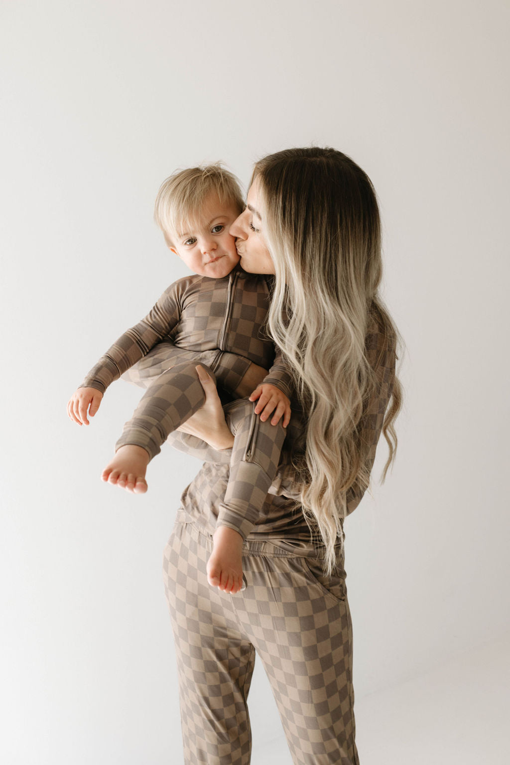 A woman with long, wavy hair kisses a toddler on the cheek. Both are dressed in matching checkered outfits from forever french baby, specifically Women's Bamboo Pajamas in Faded Brown Checkerboard, made from hypo-allergenic fabric. The background is plain and light-colored. The toddler has light hair and looks slightly towards the camera.