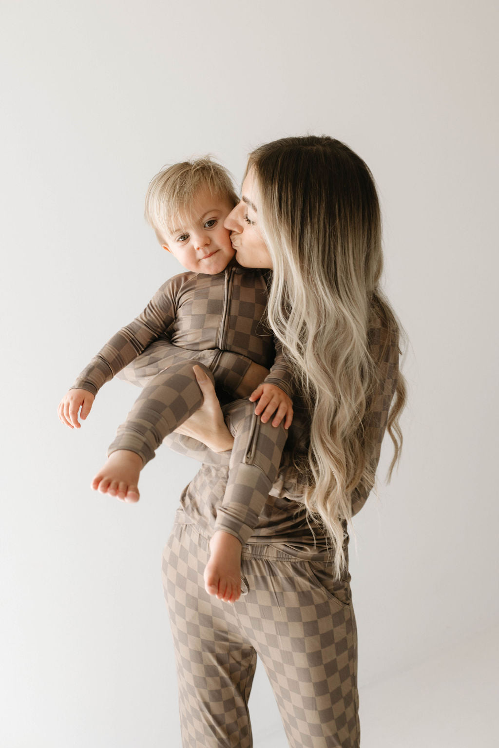A woman with long, wavy blond hair kisses a baby on the cheek. Both are wearing matching Faded Brown Checkerboard Women's Bamboo Pajamas from forever french baby, made of hypo-allergenic bamboo fabric against a plain white background. The baby is held comfortably in the woman's arms, looking content.