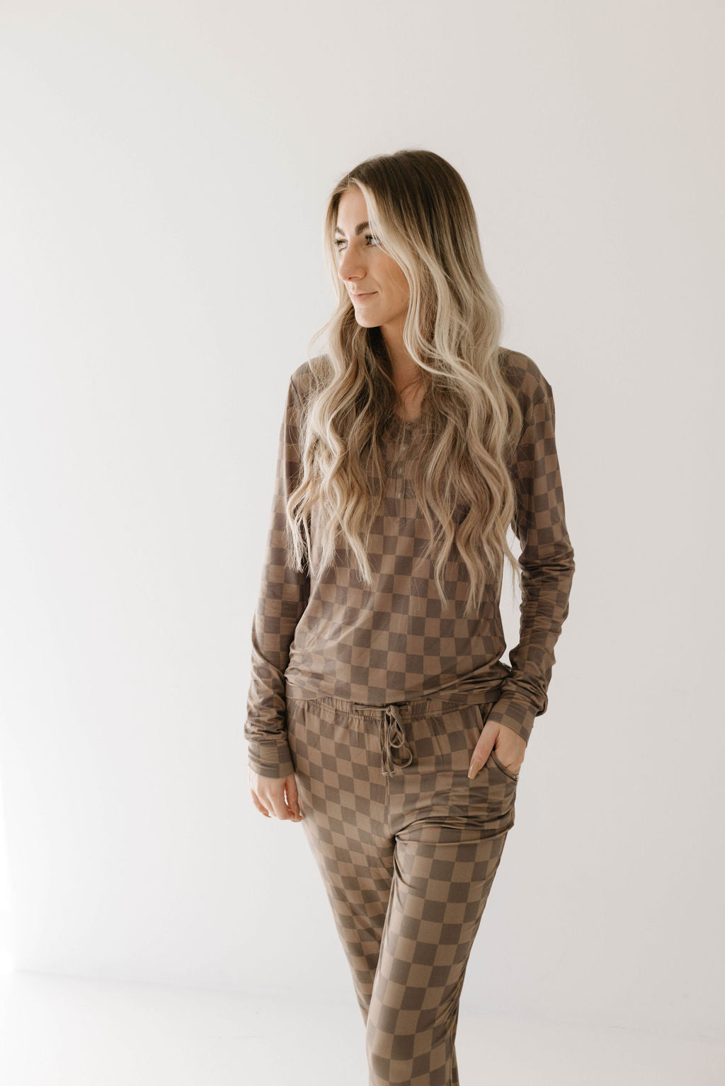 A person with long, wavy blonde hair stands against a plain background. They are wearing the Women's Bamboo Pajamas in Faded Brown Checkerboard by forever french baby, a matching hypo-allergenic bamboo loungewear set consisting of a long-sleeve top and pants in a brown and beige checkerboard pattern. Their left hand is in their pocket.