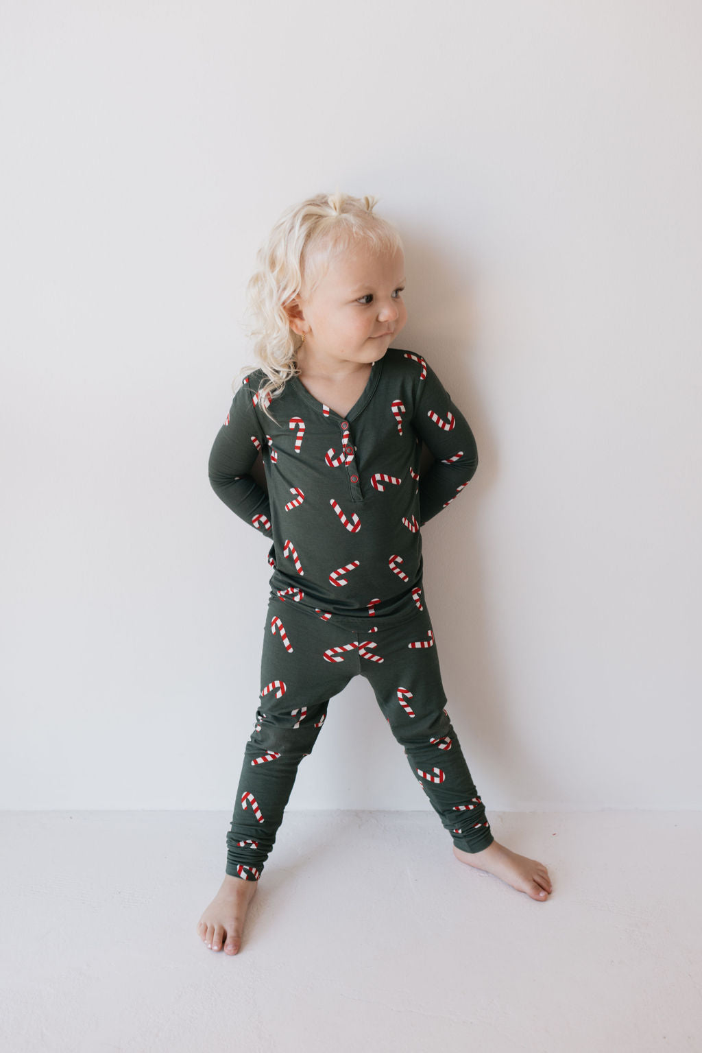 A young child with curly blonde hair stands against a light background, wearing the Bamboo Two Piece Pajamas in Candy Cane Lane design from forever french baby. The breathable and hypo-allergenic bamboo fabric ensures comfort as the child looks to the side with their hands behind their back, smiling slightly.