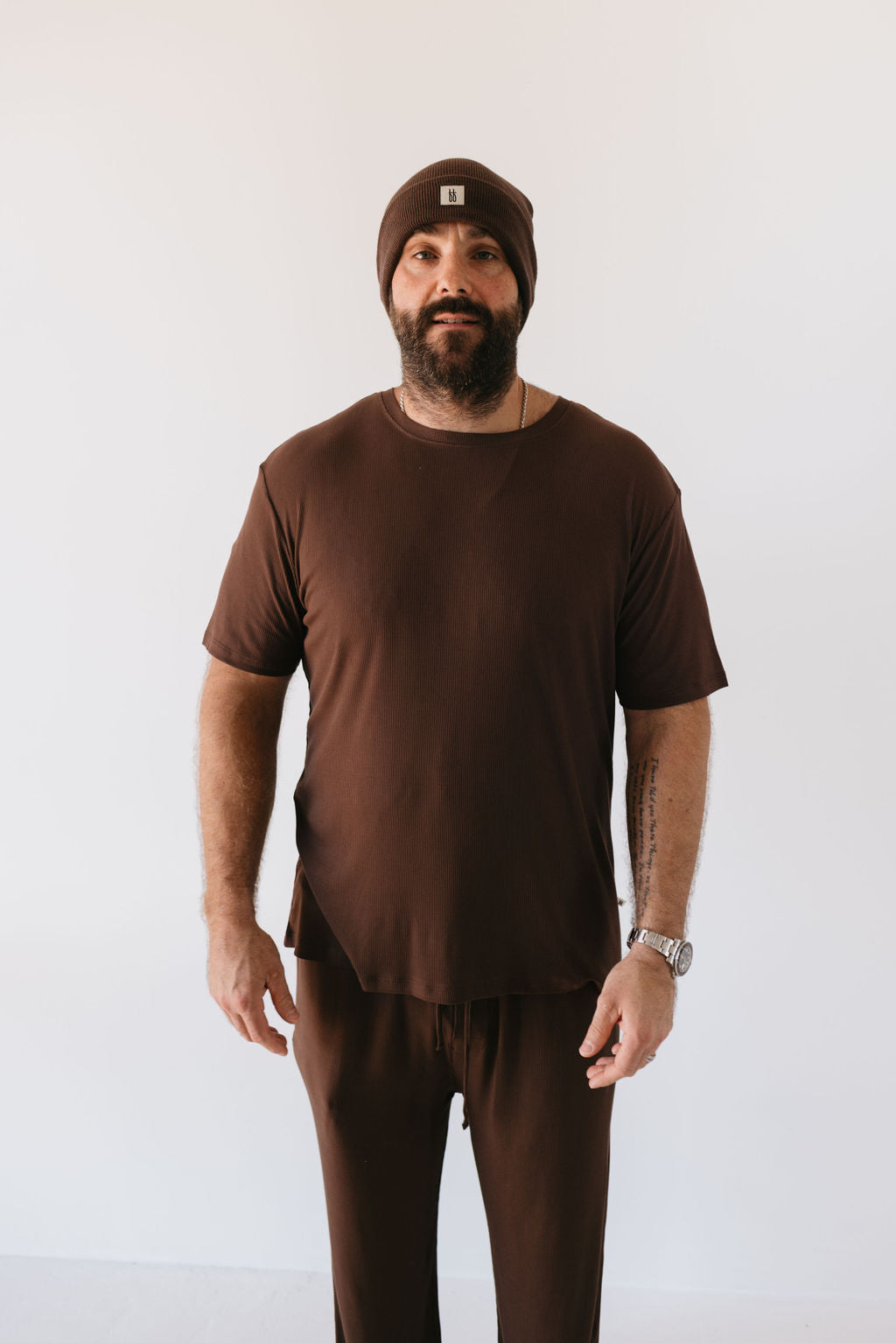 A man with a beard, donning the Men's Bamboo Short Sleeve Pajamas in Coffee Bean by forever french baby, stands against a plain white background. He has tattoos on his left forearm and wears a watch on his left wrist.