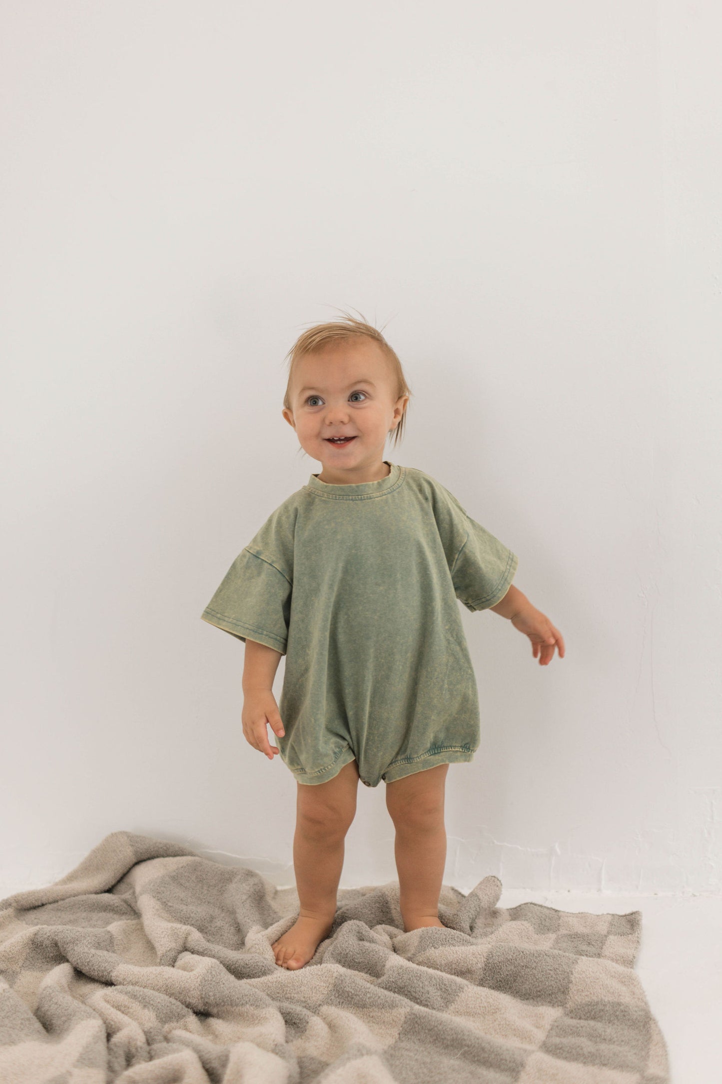A toddler with light hair stands on a forever french baby Plush Blanket in a wavy checkerboard pattern of beige and gray against a plain white background. The child is wearing an oversized, green, short-sleeve romper and is barefoot, looking slightly to the side with a small smile.