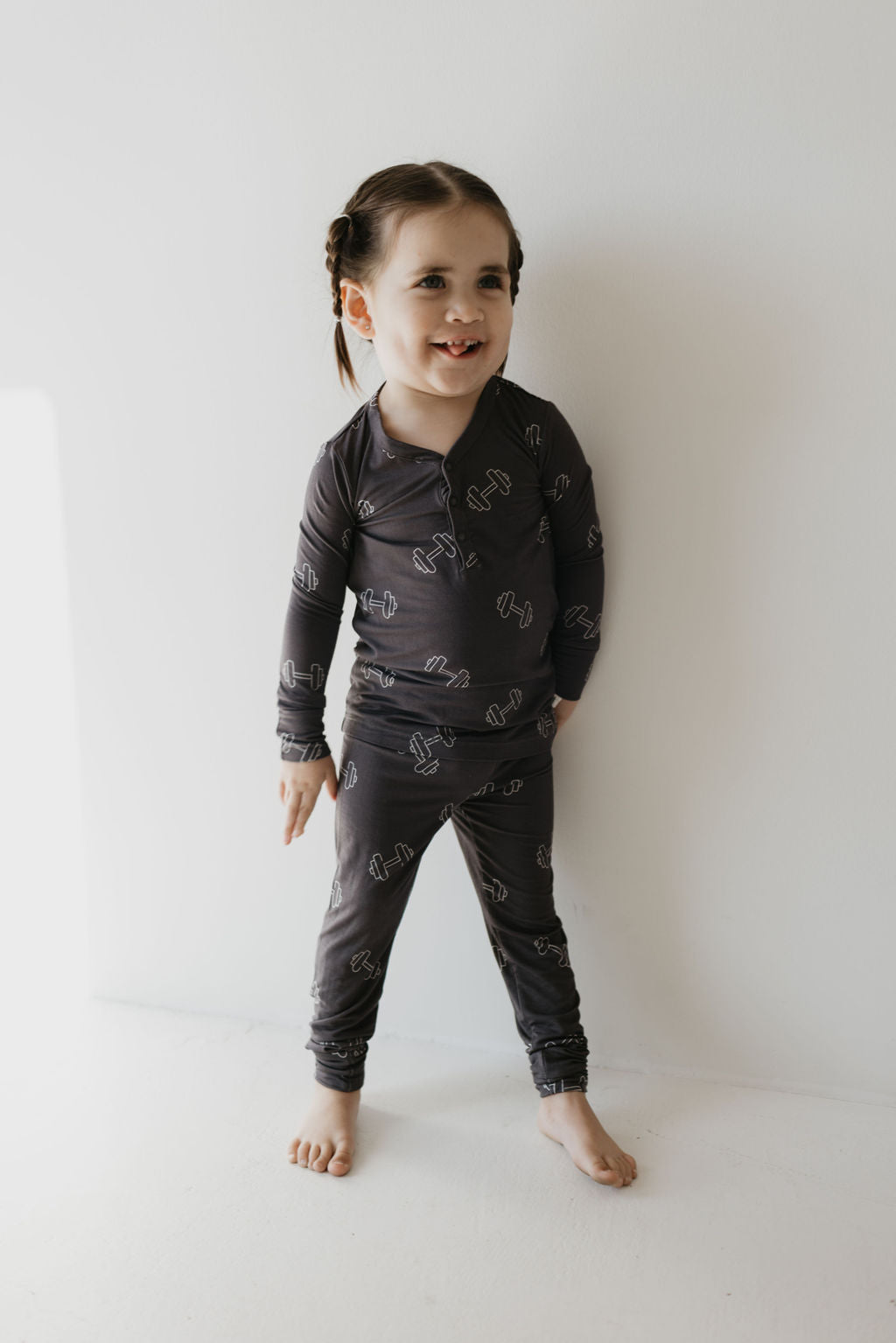 A young child with braided hair stands barefoot against a white wall, wearing forever french baby's Bamboo Two Piece Pajamas in Charcoal Arm Day. The breathable sleepwear complements their joyful expression as they smile, looking slightly to the side.