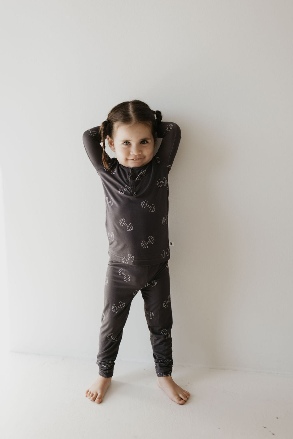 A young child stands barefoot against a white wall, smiling as they showcase their two small braids while wearing forever french baby’s Bamboo Two Piece Pajamas in Charcoal Arm Day, featuring a subtle pattern that enhances the charm of the breathable sleepwear.