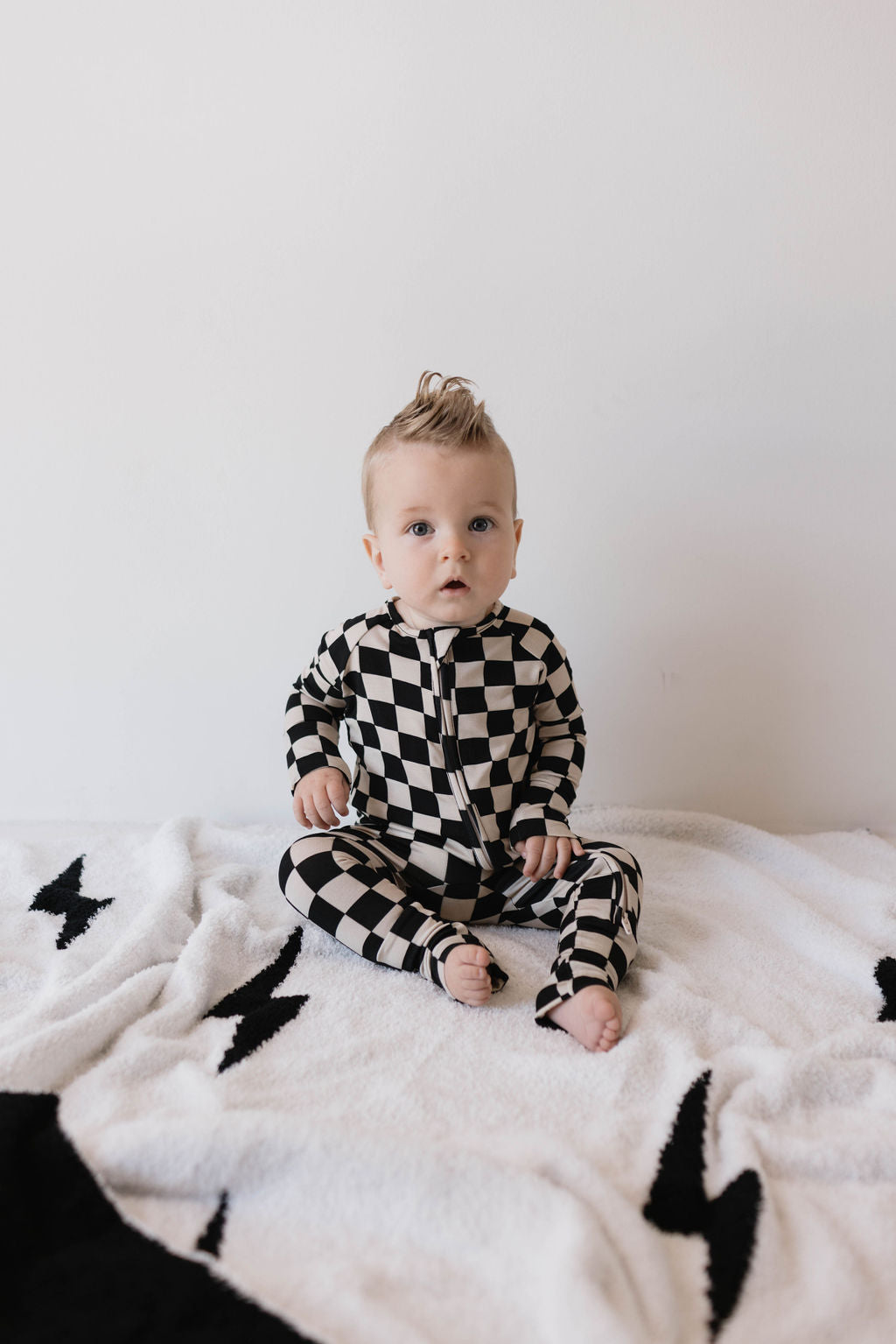 A baby with blonde hair sits on a white blanket adorned with black lightning bolt patterns. The baby, dressed in Forever French Baby Bamboo Zip Pajamas in Black Checkerboard, looks forward with a surprised expression.