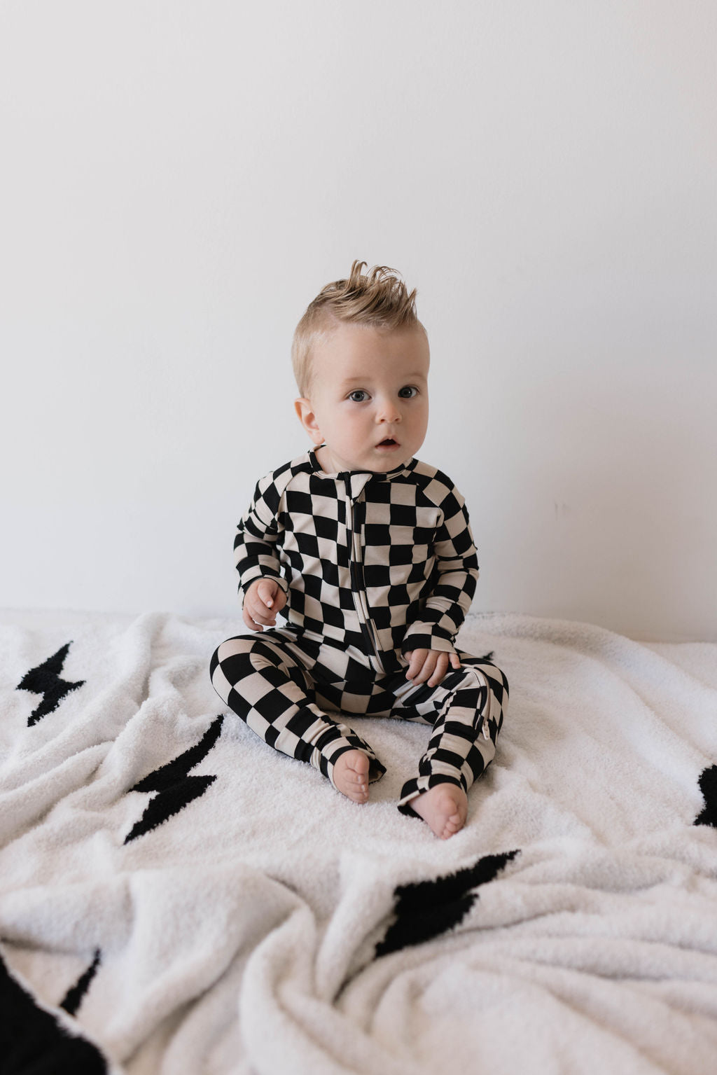 A baby with light hair sits on a soft blanket, adorned in forever french baby's Bamboo Zip Pajamas in Black Checkerboard with rollover cuffs. Against a plain white wall, the cozy blanket beneath them features charming black star patterns, highlighting the premium quality of their bamboo pajamas.