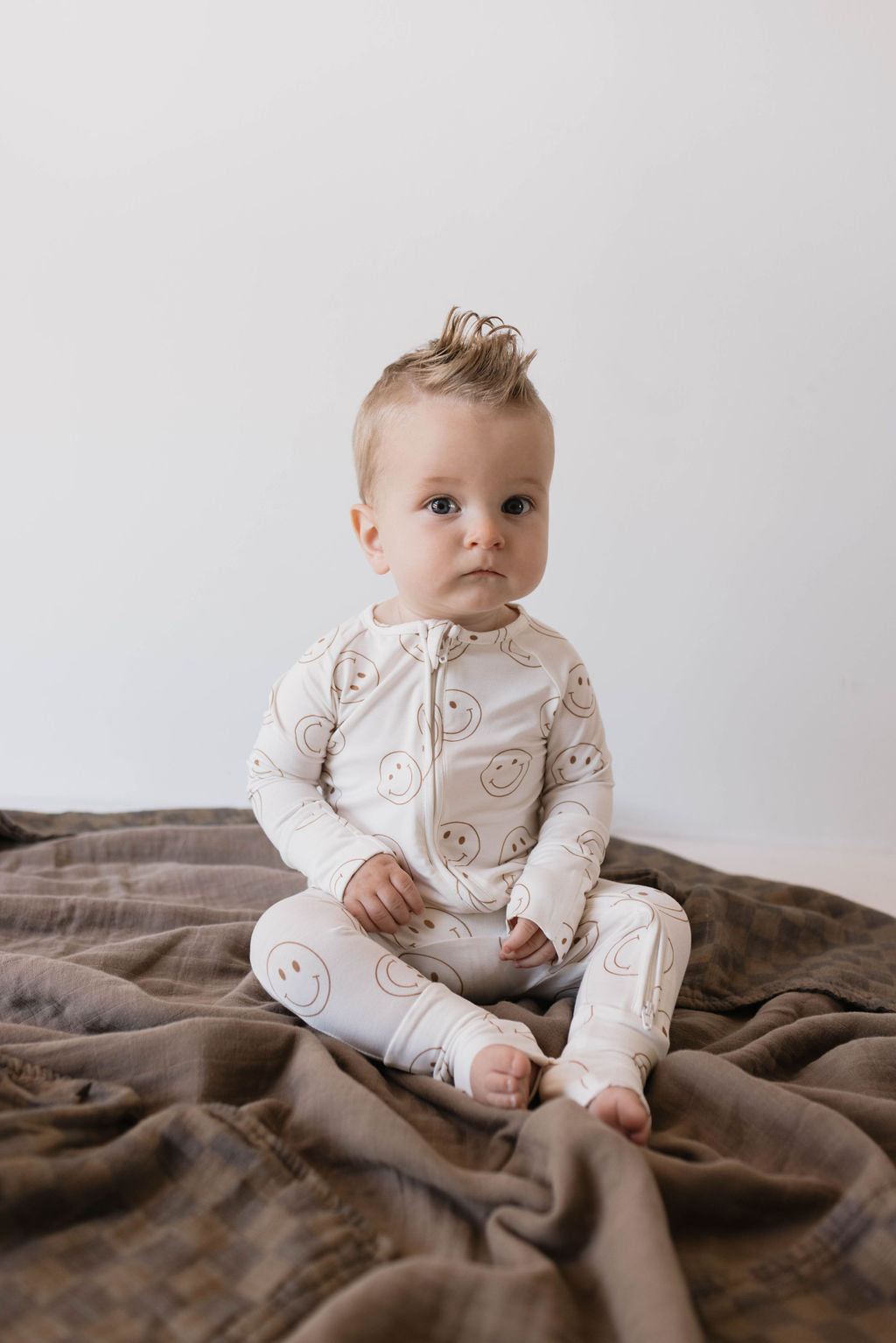 A baby with light-colored hair styled into a small mohawk is sitting on a brown blanket. They are wearing Forever French Baby's Bamboo Zip Pajamas in the 'Just Smile' pattern. The background is minimalistic with a white wall. The baby looks alert and focused.
