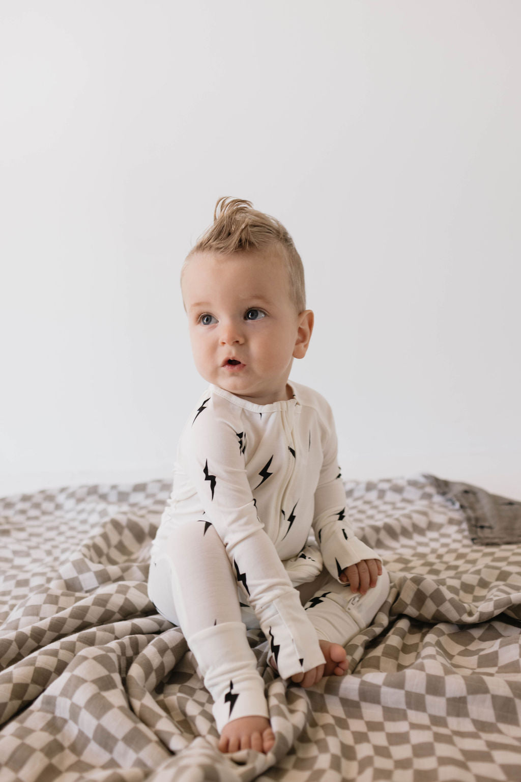 A baby with a small tuft of hair on top is sitting on a checkered blanket, wearing Bamboo Zip Pajamas | White & Black Lightning Bolt from forever french baby. The background is plain white.