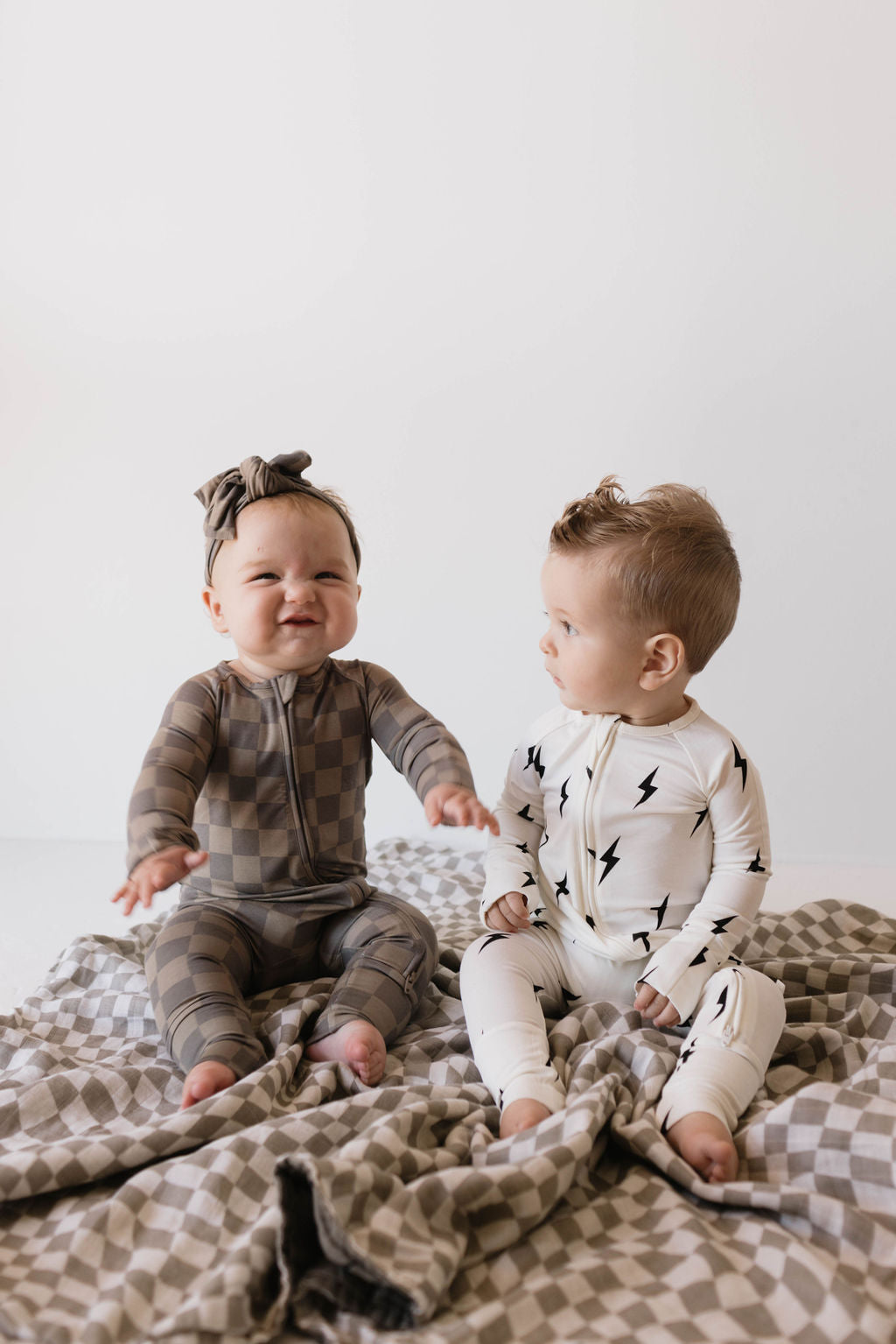 Two babies are sitting on a patterned blanket. The baby on the left wears a brown checkered onesie and a matching headband, smiling joyfully. The baby on the right sports Bamboo Zip Pajamas | White & Black Lightning Bolt from forever french baby, looking curiously at the other baby.