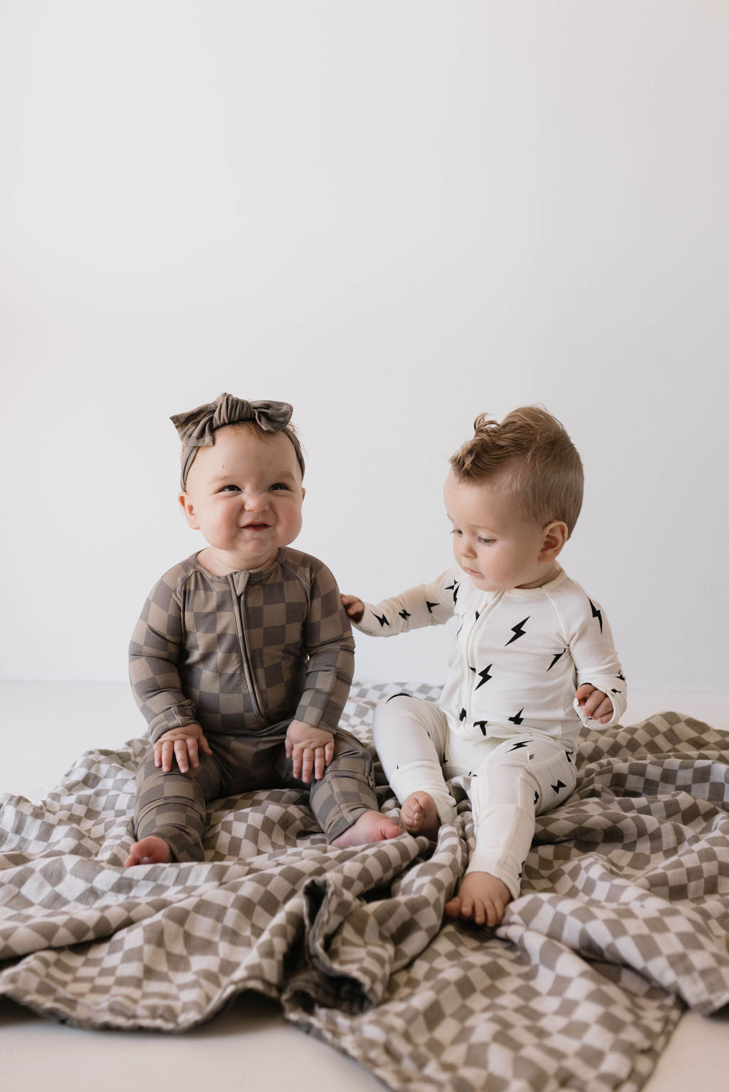 Two babies sit on a checkered blanket in a minimalistic space. The baby on the left, smiling with joy, wears the Bamboo Zip Pajamas in Faded Brown Checkerboard from forever french baby. The baby on the right, in a white hypo-allergenic onesie with black lightning bolts, reaches out towards the other baby.