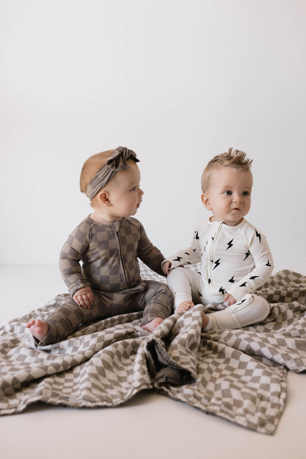 Two babies sit on a patterned blanket. The baby on the left, wearing a headband and Bamboo Zip Pajamas in Faded Brown Checkerboard from forever french baby, looks at the baby on the right, who is dressed in a breathable white onesie with black lightning bolts and looking ahead. The background is plain and light-colored.