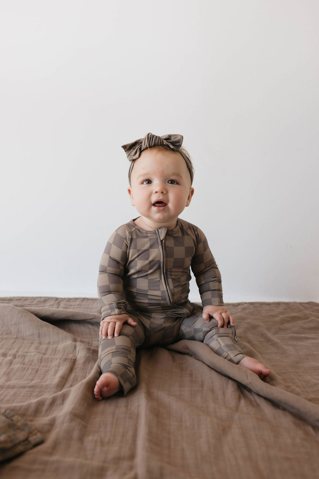 A baby dressed in a breathable, Faded Brown Checkerboard Bamboo Zip Pajamas from forever french baby, complete with a matching headband, is seated on a soft, brown blanket. The baby gazes slightly upward with a curious expression as light gently illuminates the simple, minimalist background.