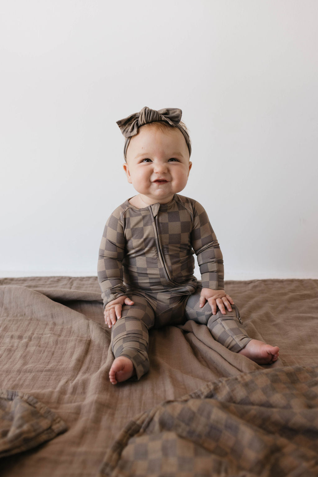 A baby wearing the "Bamboo Zip Pajamas | Faded Brown Checkerboard" by forever french baby, along with a matching headband, sits on a brown blanket. The baby has a curious expression and looks slightly upward, with their hands resting on their legs. The soft, hypo-allergenic fabric ensures comfort against the simple white background.