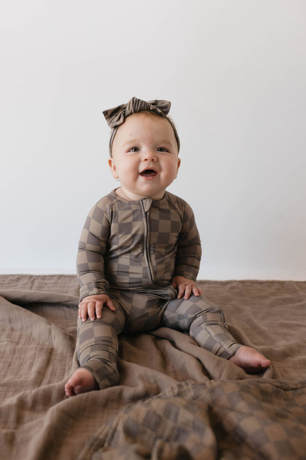 A happy baby, dressed in the "Bamboo Zip Pajamas | Faded Brown Checkerboard" by forever french baby, complete with a matching headband, sits on a beige blanket against a plain white background, smiling with joy.