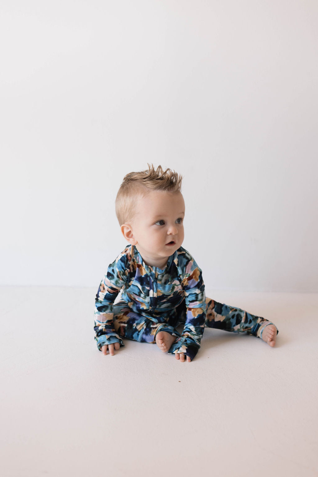 A baby with short, blonde hair and a spiky hairdo is sitting on a white floor against a plain white background. The baby is wearing colorful, patterned Bamboo Zip Pajamas in the Charli Print by forever french baby, featuring shades of blue, brown, and green. Looking slightly to the left, this adorable scene subtly promotes autism awareness.