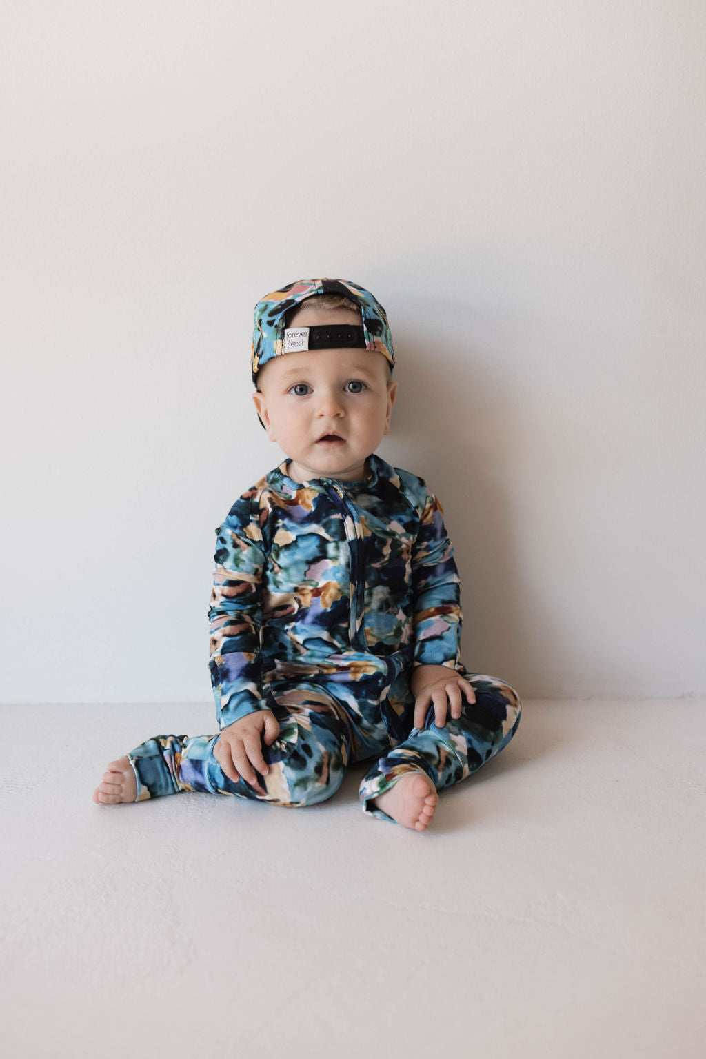 A baby is sitting on the floor against a plain white background. The baby is wearing a matching outfit consisting of a blue, black, and white camo-print hat worn backward and Bamboo Zip Pajamas | Charli Print from forever french baby with the same camo pattern. Made from soft bamboo clothing, the barefoot baby has a curious expression.