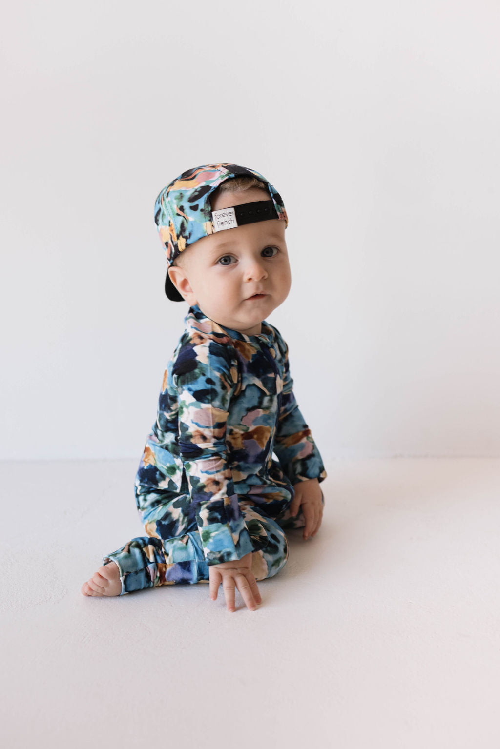 A baby sits on the floor, facing the camera but slightly turned to the side. The baby is dressed in colorful, patterned Bamboo Zip Pajamas in Charli Print by forever french baby, with a matching hat worn backwards. The background is plain and white.