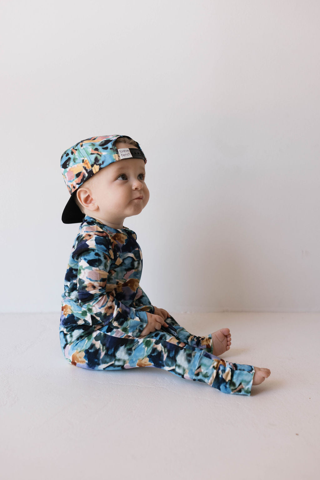 A baby with a curious expression is sitting on the floor against a plain background, wearing the Charli Print Bamboo Zip Pajamas from forever french baby. The outfit features a floral pattern in blue, green, and peach colors and includes a long-sleeve onesie and a cap worn backwards.