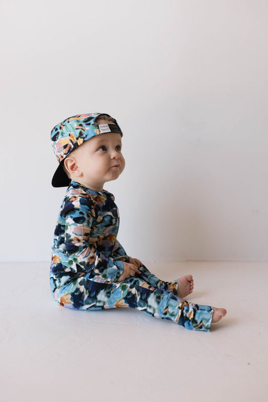 A baby with a curious expression is sitting on the floor against a plain background, wearing the Charli Print Bamboo Zip Pajamas from forever french baby. The outfit features a floral pattern in blue, green, and peach colors and includes a long-sleeve onesie and a cap worn backwards.